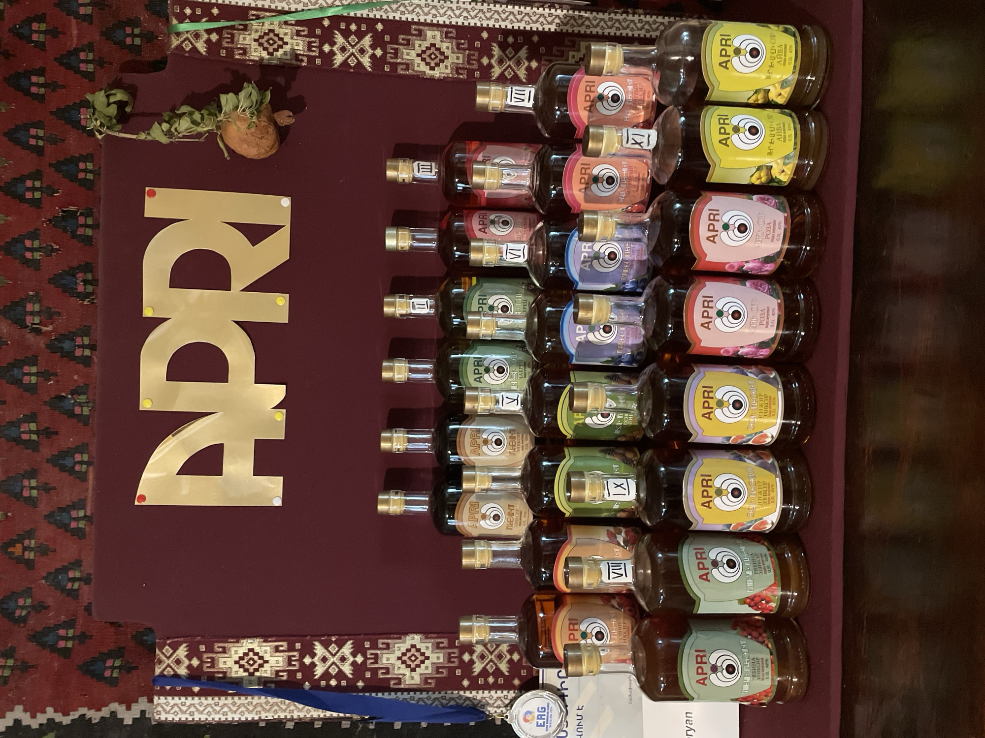 The image depicts a collection of alcoholic beverages, likely brandies or fruit liqueurs, displayed in glass bottles with gold caps. The bottles are arranged in multiple rows on a dark red surface, with the brand "APRI" prominently featured on their labels, often accompanied by imagery of fruits or flowers. Some bottles have Roman numerals on their labels, suggesting different varieties or ages. Behind the bottles, a large, gold, three-dimensional "APRI" logo is affixed to a dark red backdrop. To the left of the logo, a medal with an "ERG" inscription is partially visible, attached to a blue ribbon. The background features a patterned textile, likely a rug, with geometric designs in red, black, and green. A dried fruit or vegetable with wilting leaves hangs from a string to the right of the "APRI" logo. The scene is indoors, with ambient lighting. The location is identified as Stepanavan, Armenia.