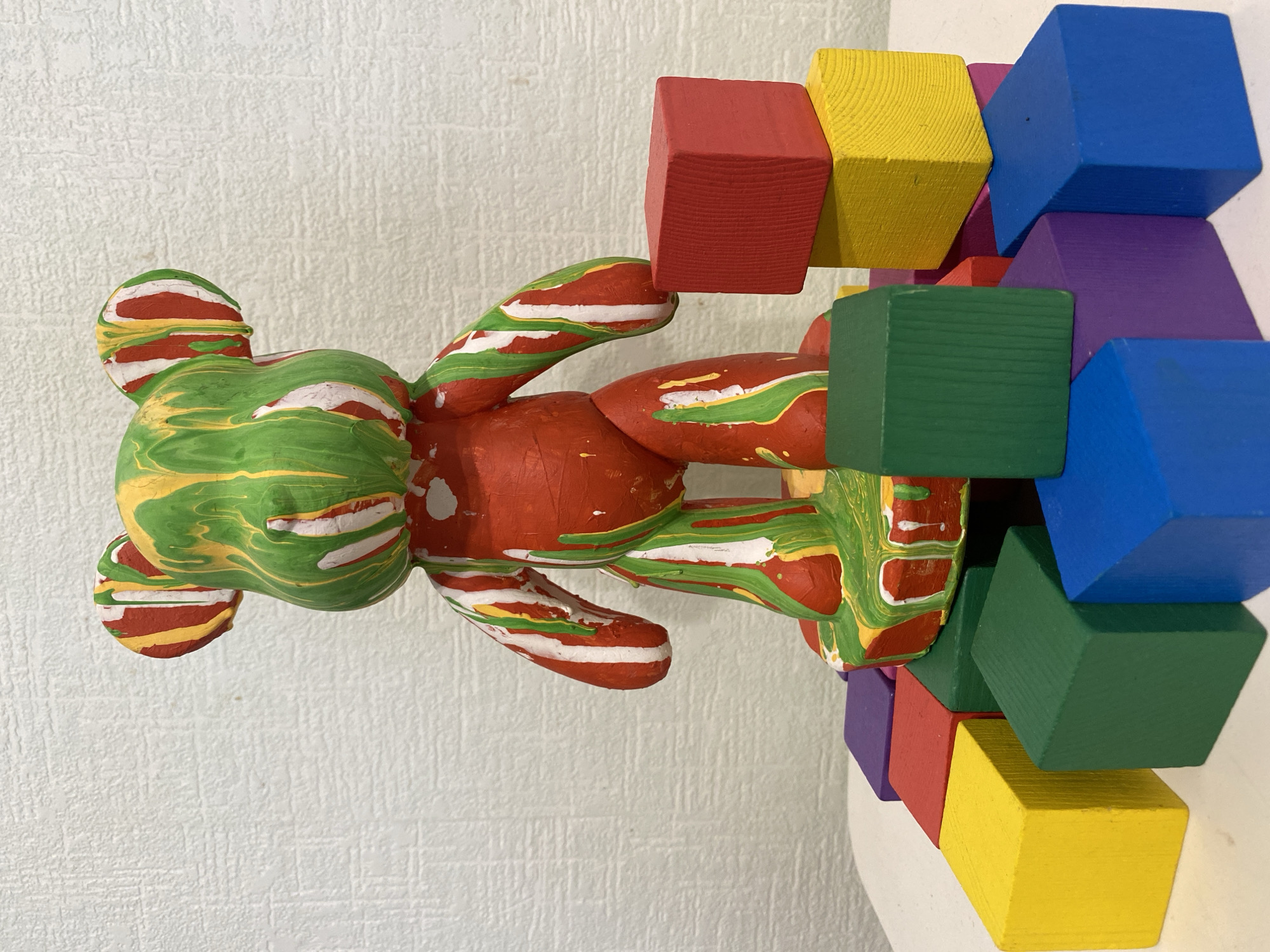 The image shows a painted bear figurine propped against a stack of wooden blocks. The bear is predominantly green and red with white and yellow accents. The wooden blocks are various colors, including red, yellow, blue, green, and purple. The background is a textured off-white wall. There is no indication of a location, or the city of Sochi, Russia.