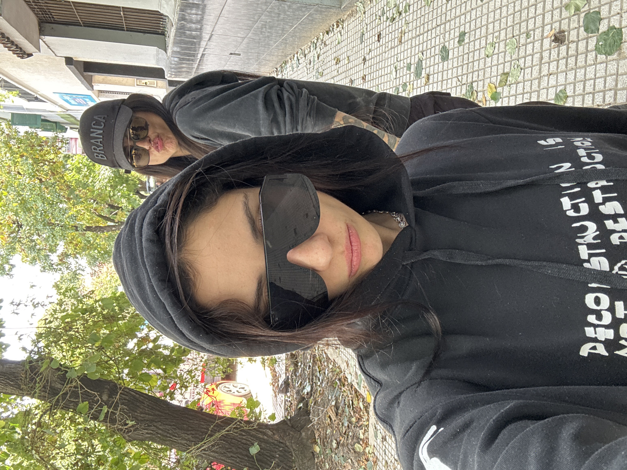 The image is a selfie of two individuals, presumed to be women, positioned on a sidewalk in Buenos Aires, Argentina. The primary subject is in the foreground wearing a black hooded sweatshirt and large black sunglasses. The hoodie has white text, partially visible, with letters spelling out "DECO" and the partial phrase "BEST ACTION". A silver necklace is visible around the primary subject's neck. The secondary subject, slightly out of focus, wears a black cap with white lettering "BRANCA" and sunglasses. Both subjects are standing outside on a sidewalk. The sidewalk is a patterned stone pavement covered with fallen leaves. A street borders the sidewalk, and beyond it is a tree with green leaves. Buildings are visible in the background.