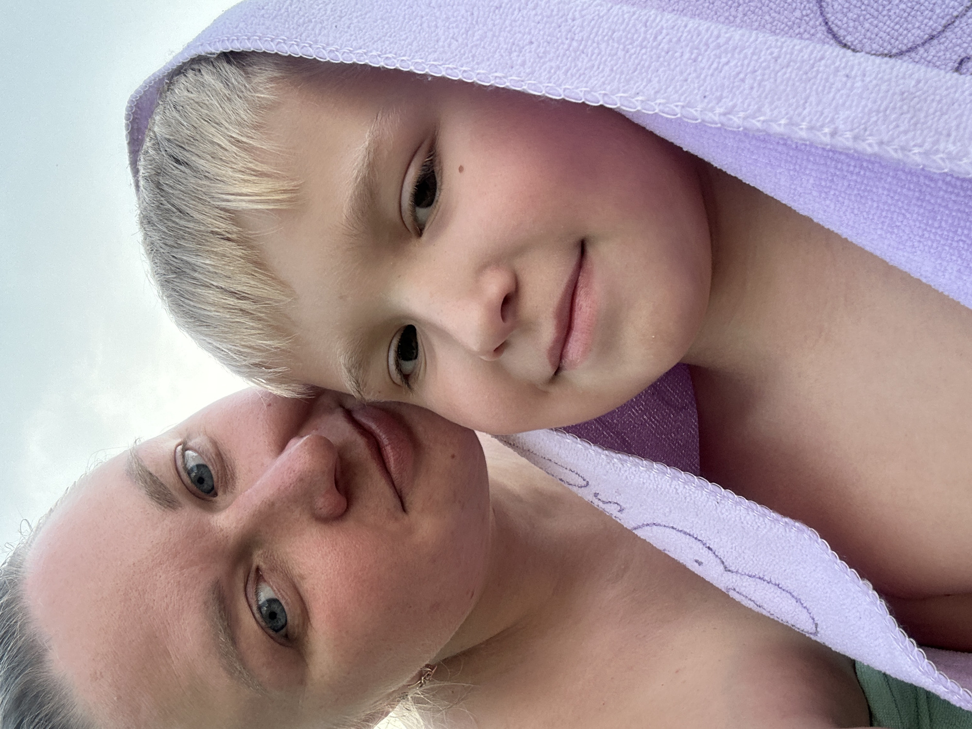 A selfie taken outdoors shows a woman and a young boy wrapped in a light purple towel. The woman is to the left, her head tilted slightly, looking towards the camera with blue eyes and light blonde hair pulled back. The boy is in the foreground to the right, with blonde hair peeking out from under the towel, smiling at the camera. The towel has a faint embroidered design. The background is a pale, overcast sky. The location is identified as Chaikovsky, Russia.