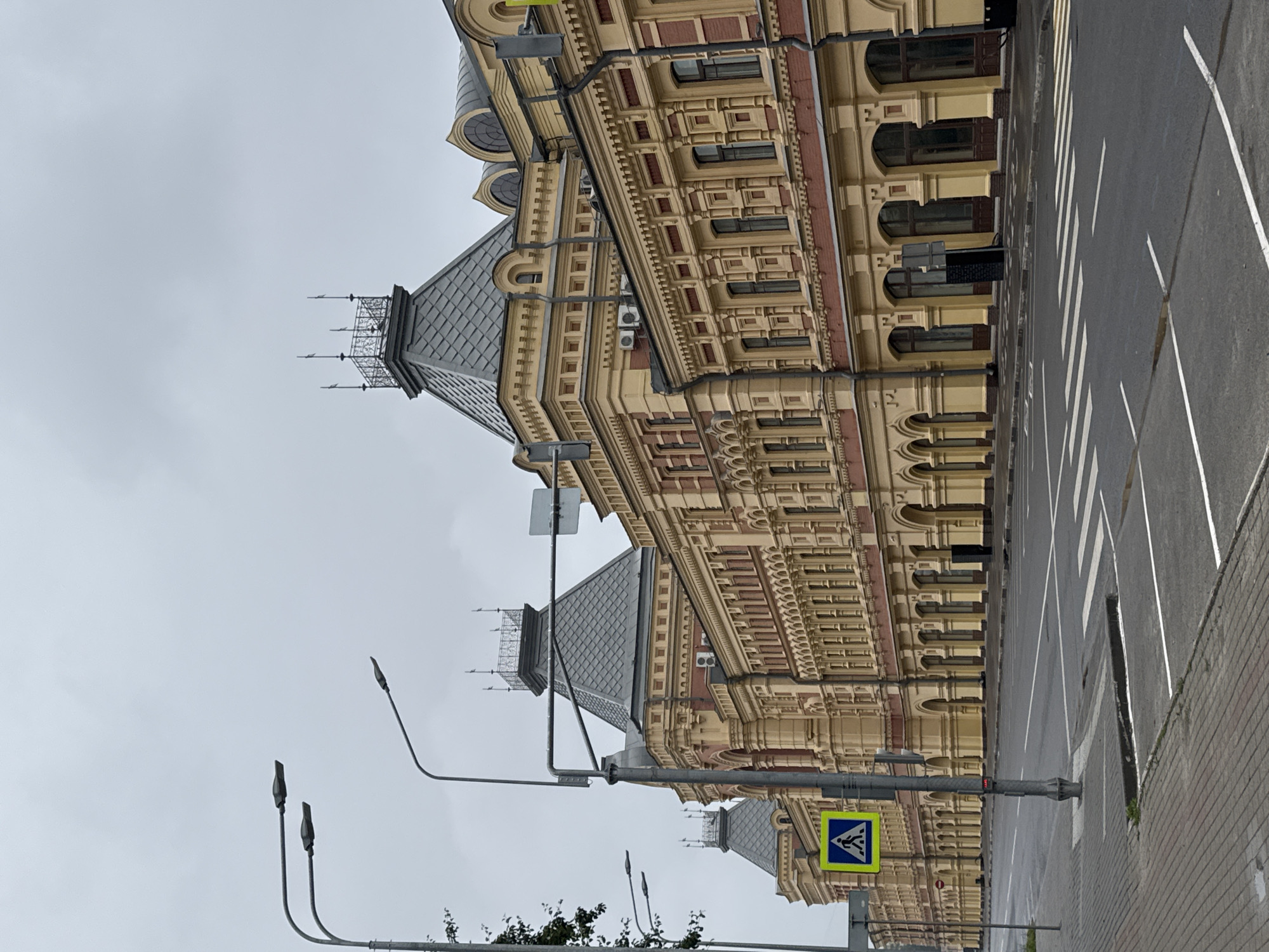 A street scene in Nizhny Novgorod, Russia, features a grand, historic building with yellow and red brick facade, arched windows, and ornate architectural details. The building, likely a government or public institution, is situated along a wide, empty street with a pedestrian crossing. Streetlights and traffic signals are visible, suggesting an urban environment. The sky is overcast, indicating a cloudy day. The image captures a sense of architectural grandeur and a quiet, orderly streetscape.