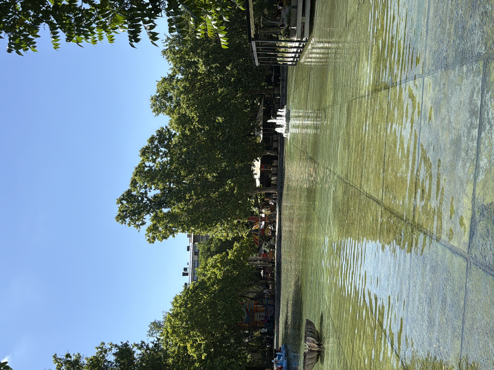 The image depicts a park scene in Yerevan, Armenia. It features a shallow body of water with algae growth, reflecting sunlight and creating ripple patterns. The water's edge is defined by concrete slabs. Trees line the left side of the frame, casting shadows on the ground. In the background, blurred figures of people are visible near a carousel and a building. A low metal railing is visible along the right side, near where the water features spray small fountains.