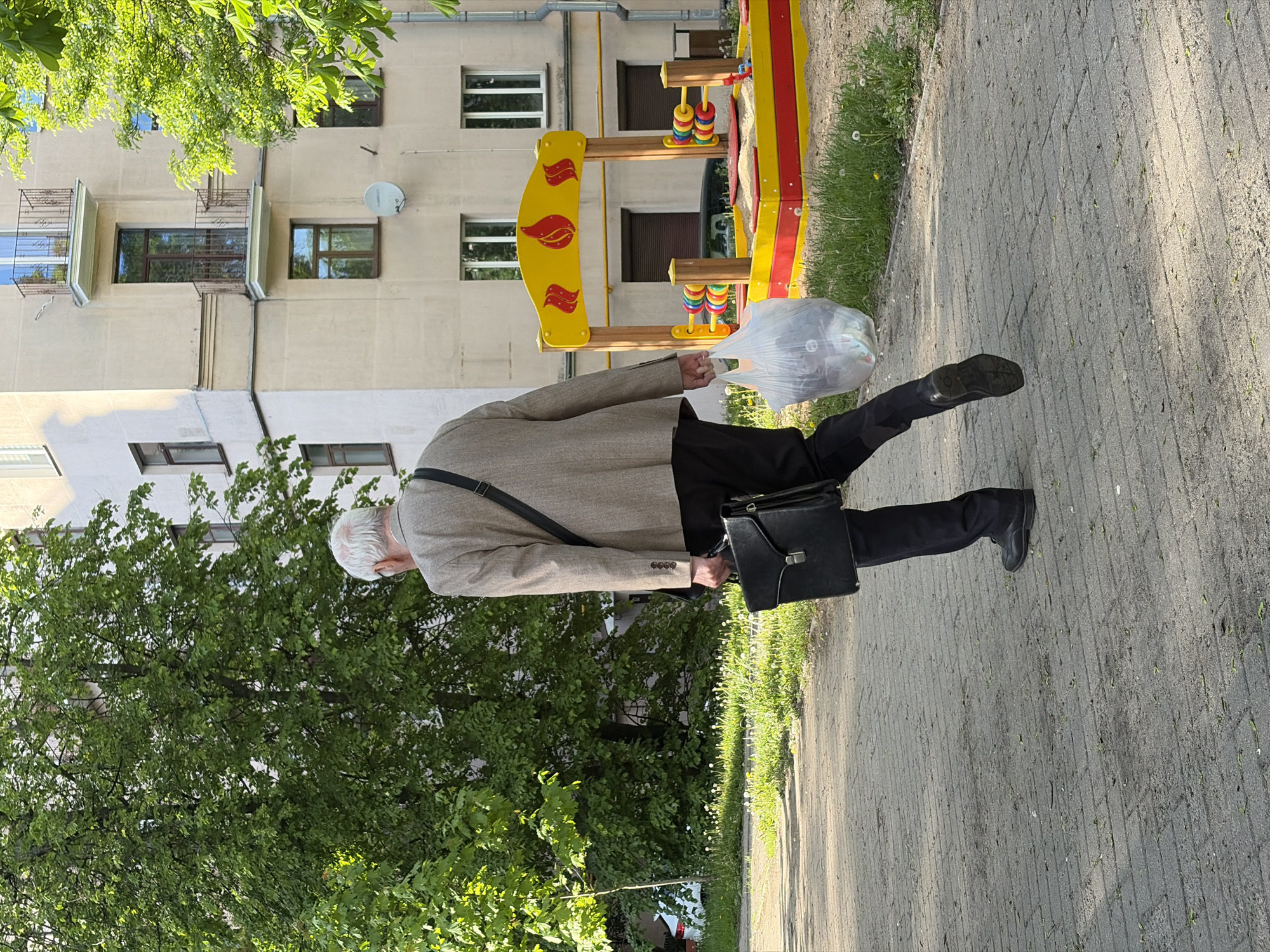 An elderly Caucasian male with white hair is walking on a paved sidewalk in what appears to be a residential area in Minsk, Belarus. He is wearing a beige jacket, black trousers, and black shoes. He carries a black briefcase on his left side and a clear plastic bag in his right hand. The bag seems to contain multiple objects. The man also has a strap across his chest. To his left is a tree. In the background, there is a residential building and a yellow and red playground structure. The pavement shows signs of wear and scattered debris. Sunlight is present, casting shadows in the scene.