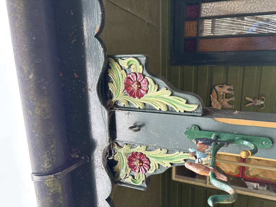 This image captures a forensic view of an exterior building structure in Stiperstones, United Kingdom, specifically focusing on a section of a dark green facade. A dark, moss-covered gutter system with a visible support bracket spans the top. Below the gutter, a dark green scalloped eave overhangs a prominent dark green vertical wooden beam. Attached to this beam are two ornate carved wooden corbels, each depicting a red flower with a yellow center encircled by light green leaves, set against a dark green background. Cobwebs are evident around these decorative elements and under the eave. A small, dark metal ring is secured to the beam between the corbels. Further down the beam, a dark green metal coat hanger with three hooks is mounted. Perched on the upper hook are three small, carved wooden birds: one brown, one blue, and one black, positioned in close proximity. To the right of the beam, on the dark green wall paneling, two wooden decorative items are affixed: an intricately carved elephant with colorful detailing above, and a smaller carved rabbit head with red floral accents below. A window with a dark frame is partially visible on the far right, featuring multi-colored, likely stained, glass panes (orange, amber, clear) in its upper section, and a reflective lower pane showing a distorted reflection of a light blue garment. The overall structure displays weathering, including moss, lichen, and rust-colored marks. No individuals are directly present.
