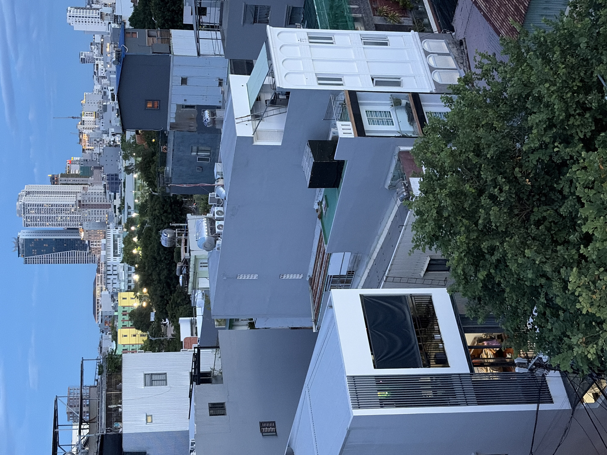 The image depicts an urban landscape in Đà Nẵng, Vietnam, at dusk. The foreground is dominated by a modern, multi-story building with white vertical louvers and a dark awning covering a balcony or window. To the right, a dense canopy of green trees partially obscures other buildings.
In the mid-ground, several buildings of varying heights and architectural styles are visible. There are structures with flat roofs, some painted in gray tones, others in white. One building features a blue roof and a distinctive orange light emanating from a window. Another prominent building is white with arched windows on the lower levels and multiple rectangular windows on upper floors. Air conditioning units are visible on the roofs of some buildings, and a water tank is mounted on one.
The background reveals the city's skyline with taller skyscrapers, including two distinctive, modern towers with angled facades, characteristic of contemporary urban development. The sky is a soft blue, indicating the transition from day to night, with lights beginning to illuminate some of the buildings. The overall scene conveys a sense of a densely populated, developing city.
