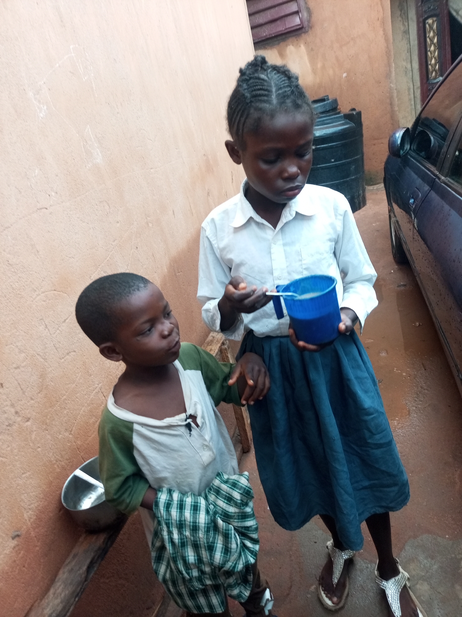 The image depicts two young children standing outdoors in what appears to be an urban environment in Tempure, Nigeria. A young girl, dressed in a white collared shirt and a dark blue skirt, holds a blue mug with a spoon in her right hand, appearing to offer a drink to a younger child. The younger child, wearing a green and white long-sleeved shirt, stands to the left, looking towards the mug. A metal bowl is visible on a wooden surface near the younger child. In the background, a large black water tank and a dark-colored vehicle are present. The wall behind the children is an earthy-toned stucco. The ground is a reddish-brown, wet surface, suggesting recent rain.