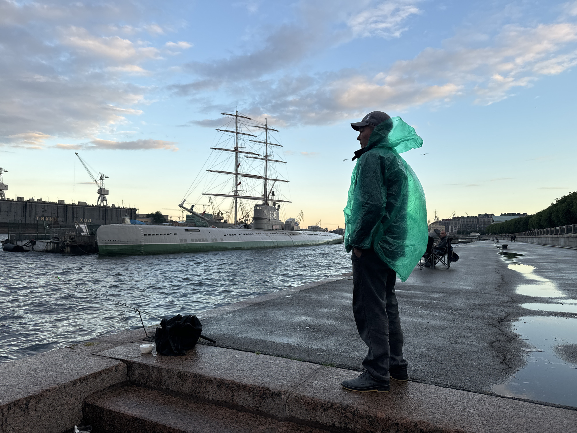 A man wearing a green poncho and a baseball cap stands on a granite embankment, facing away from the camera and towards a body of water. A fishing rod is set up on the embankment to his left. In the background, a large submarine and a sailing ship are docked near industrial cranes. The sky is partly cloudy with hues of blue and white. The embankment is wet, with puddles on the asphalt walkway. The scene appears to be a port in Saint Petersburg, Russia, possibly near the Neva River.