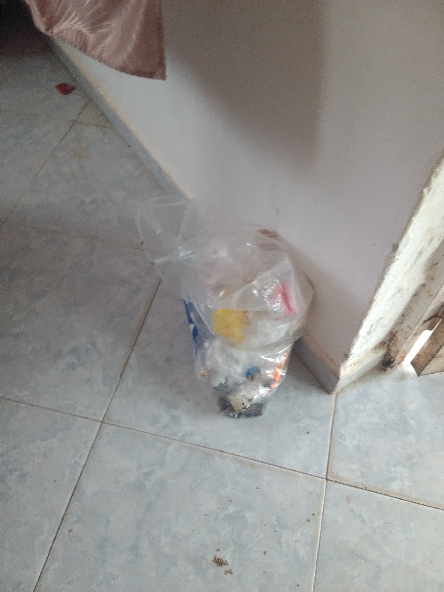 The image depicts a corner of a room with light blue tiled flooring. A clear plastic bag, partially filled with various items and some debris, is placed in the corner where two walls meet. The contents of the bag are difficult to discern clearly due to the transparency of the plastic and the focus of the image, but some colored items, possibly packaging or small objects, are visible within. To the left of the bag, near the wall, is a small red object on the floor, and a brown fabric drape is visible hanging from above. The walls appear to be painted white, and the corner where the bag sits shows some wear or dirt. The lighting suggests an indoor setting, possibly during the day due to the ambient light. No people are visible in the image.