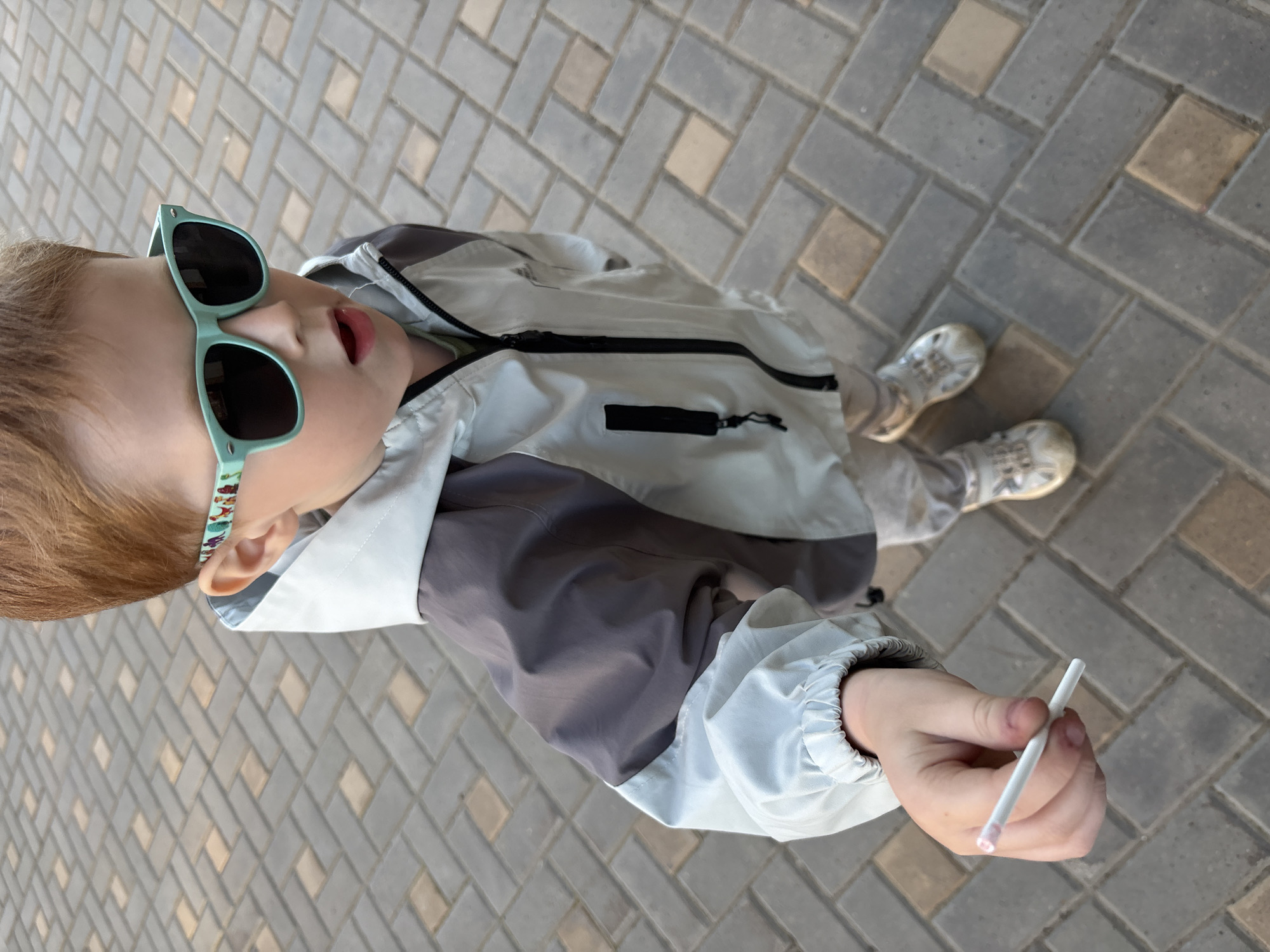 A young child, viewed from above, stands on a paved surface in Izhevsk, Russia. The paving consists of rectangular blocks laid in a staggered pattern, colored in shades of gray and beige. The child wears light-colored athletic shoes, light gray pants, and a light gray jacket with dark gray sleeves and a black zipper. They also sport sunglasses with light blue frames that are decorated with small figures. The child holds a thin white stick in their right hand. Their mouth is slightly open. The image provides no further information to confirm the city and country.