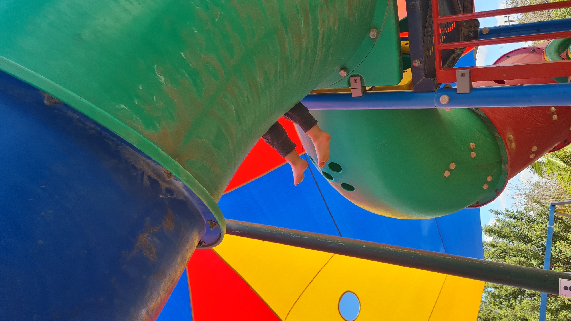 A low-angle shot shows a brightly colored playground structure with blue, green, red, and yellow components. A pair of feet, clad in grey pants, dangles from inside a green tube-like section of the structure. The location is likely Lachish Regional Council, Israel.