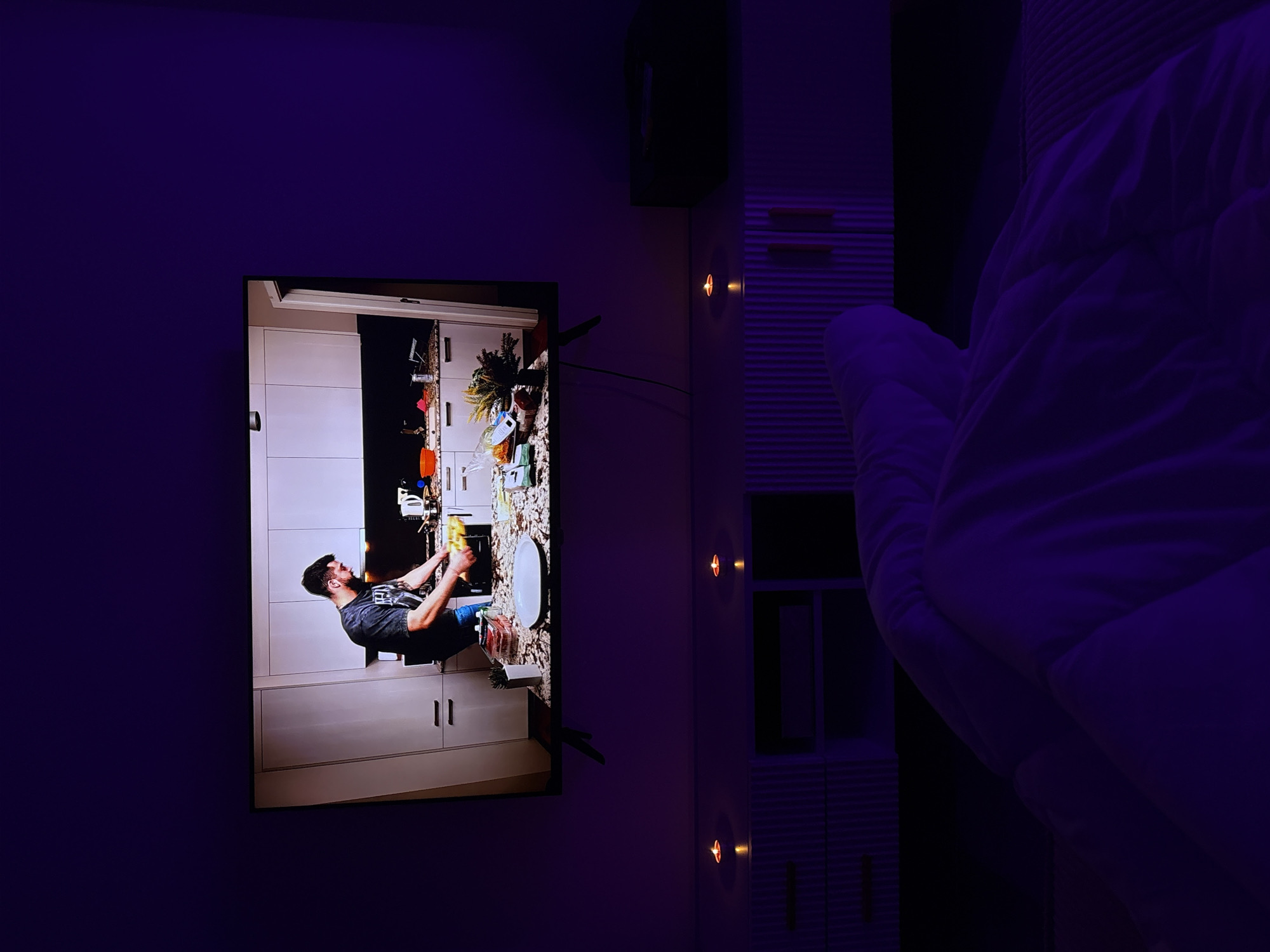 The image shows a television screen displaying a man in a kitchen, likely doing dishes. The TV is mounted on a wall in a room with a purple hue. To the right of the TV is a shelving unit, and a portion of a puffy, white blanket or comforter is visible in the foreground. There are a couple of sconce-like lights on the wall. The image could have been taken in Mönchengladbach, Germany, but there's no visual confirmation within the image.