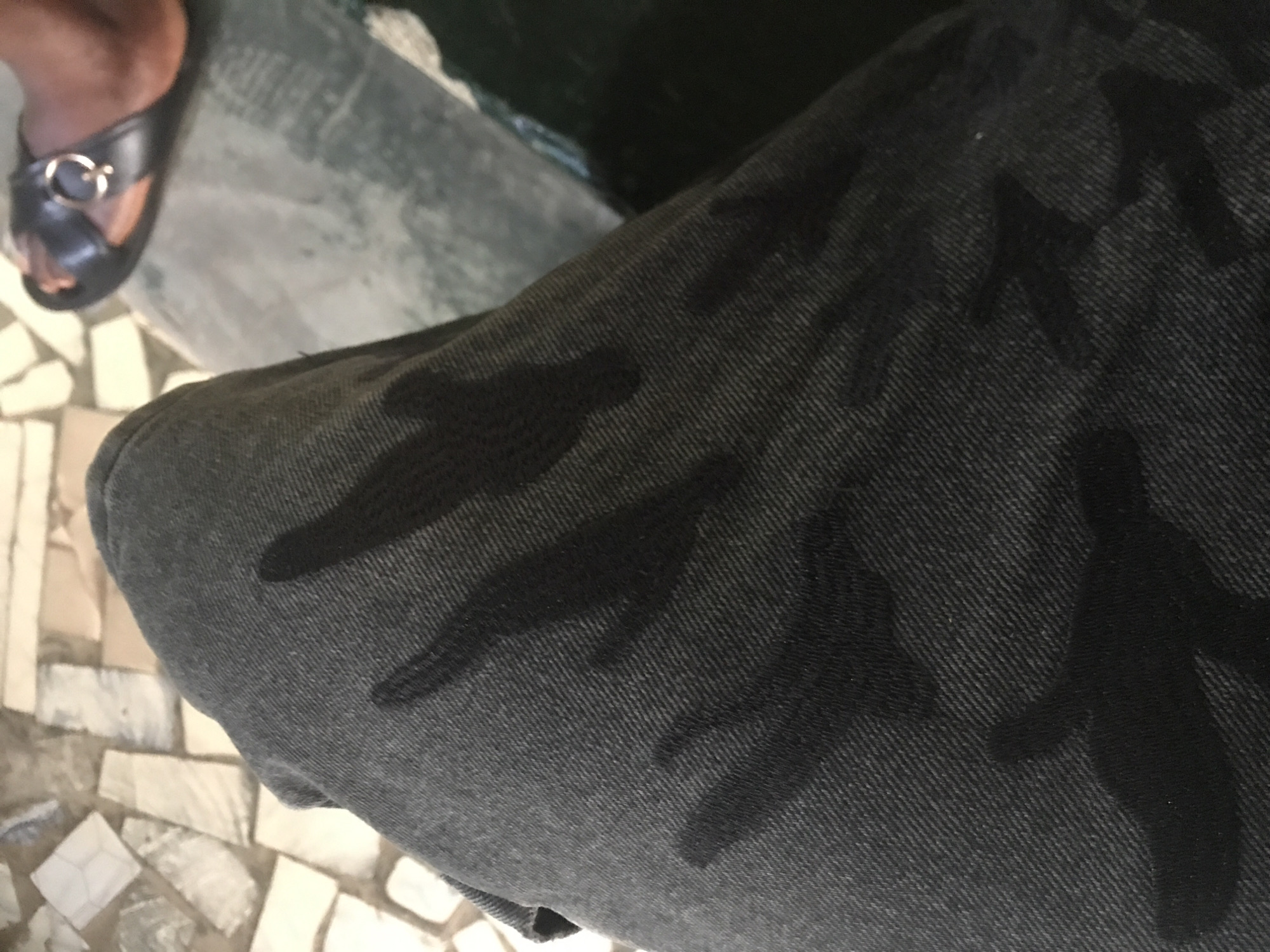A close-up, overhead shot shows a person's dark grey denim pants with wet patches. The wet areas are darker than the rest of the fabric, indicating moisture. The pants appear to be resting on a tiled surface. In the upper right corner, a black sandal with a gold buckle is visible on a person's foot, suggesting they are wearing the pants. The background is out of focus but appears to be an outdoor setting with similar tiling and possibly some green foliage.