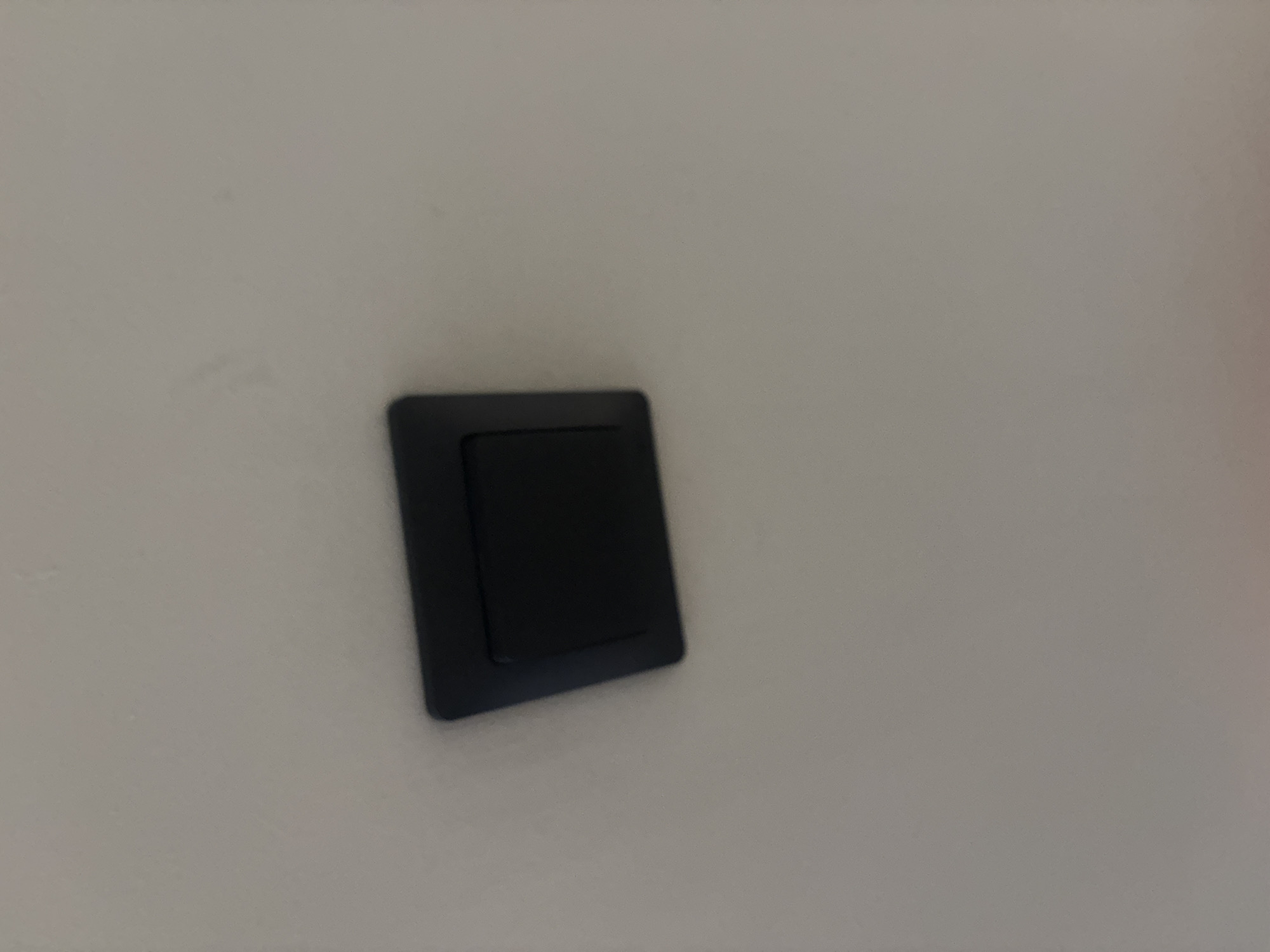 A close-up, high-angle shot shows a black electrical wall plate with a single toggle switch mounted on a light-colored ceiling. The plate is square with beveled edges. The switch is in the center of the plate and appears to be in the "off" position. The ceiling is textured and shows some subtle variations in shade, possibly from light or imperfections. The image lacks any contextual information about the location within the city of Ldzaa, Abkhazia, or any details about people or actions.