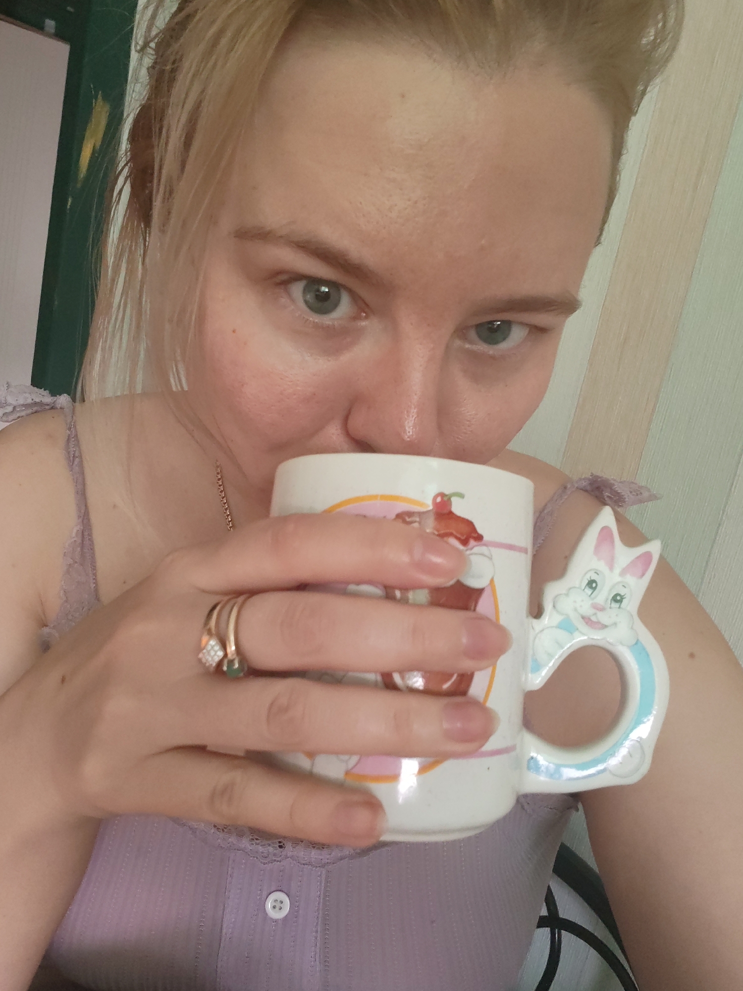 A woman with blonde hair, fair skin, and visible facial pores holds a white mug with a colorful milkshake illustration and a bunny-shaped handle near her mouth. She wears a light purple lace-trimmed top and a gold chain necklace. Two gold rings are visible on her left hand. The background shows a light green and white striped wall on the right, and a dark green door frame on the left. The location appears to be indoors, with no indication of being in the city of Izhevsk, Russia.