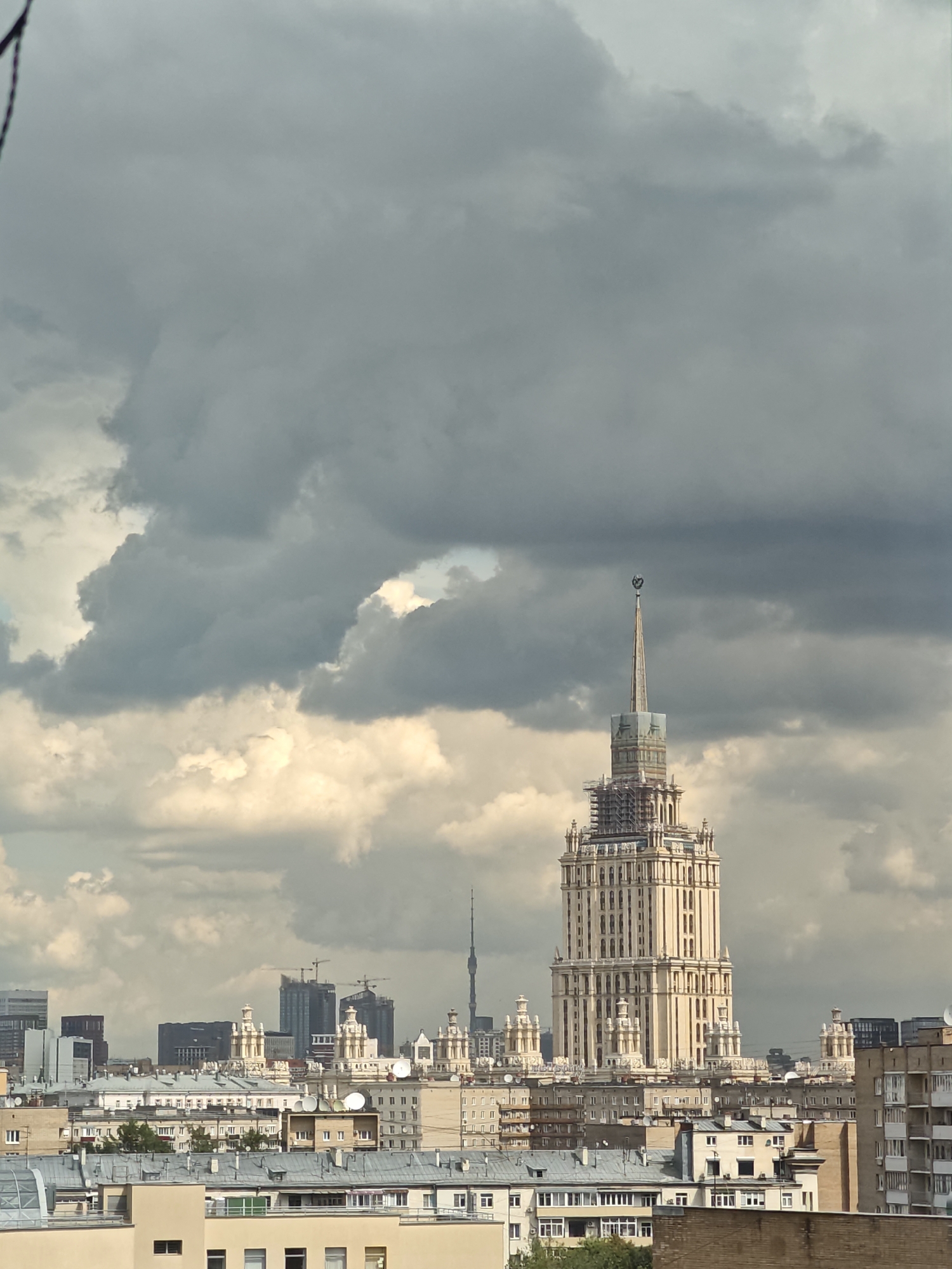 A high-angle, full shot captures a city skyline under a dramatic, cloudy sky. Dominating the foreground is a tall, ornate, light-colored skyscraper with a pointed spire topped with a star. The building appears to be undergoing renovation, with scaffolding visible around its upper sections. To the left and right of the main skyscraper, other buildings of varying heights and architectural styles form the urban landscape. In the distance, a slender television tower is visible on the left. The sky is filled with a mix of dark, heavy storm clouds and lighter, illuminated clouds, creating a sense of foreboding or an approaching storm. The overall scene is a view of a dense urban environment with a prominent landmark building against a turbulent sky.