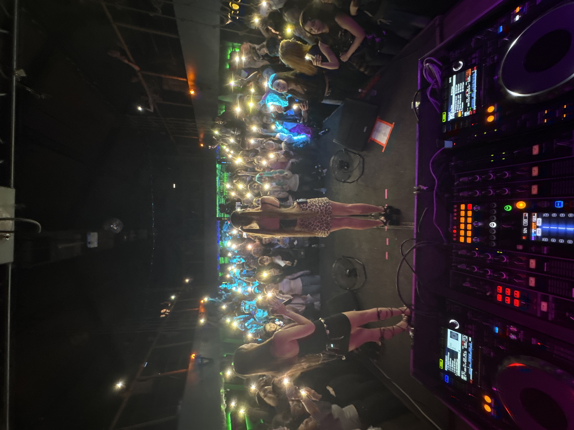 This image depicts a vibrant indoor music event, likely a club night or concert, taking place in Chita, Russia. The setting is dark, predominantly lit by artificial lights from the stage and the audience's smartphones.
In the foreground to the right, a professional DJ setup is visible, featuring turntables and a mixer with multiple glowing buttons and digital screens. These screens display technical information such as "01:02.29.5", "0.00.157", "6511105", "158.9", "CH2 CH3", and "03.25.26.5.-0.05. 158", along with letters "C", "D", and "CCC", indicating track times, channels, or other playback details. A hand is seen adjusting controls on the mixer.
Two women are on a stage or platform to the left of the DJ equipment. One woman, with long, light hair, is seen from behind, wearing a leopard print skirt and facing the audience. Another woman with long dark hair is also on the stage, wearing a dark outfit and appearing to interact with the crowd. A black fan is positioned on the stage floor near them.
A large crowd fills the rest of the venue, extending into the background. Many attendees are actively participating by holding up their smartphones with flashlights activated, creating a dense pattern of bright white and green lights that illuminate parts of the audience. The stage itself is bathed in green, blue, and purple lighting, contributing to the energetic atmosphere of the event.