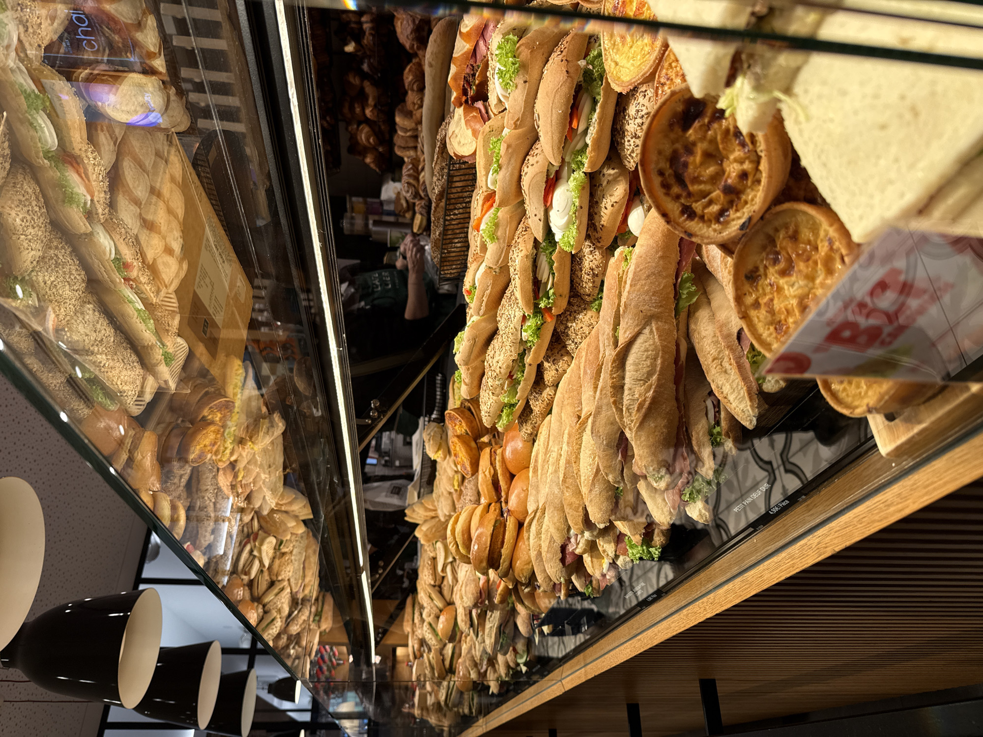 The image shows a display case filled with various food items, seemingly within a bakery or delicatessen in Luxembourg, Luxembourg.
The primary contents of the display case are sandwiches, pastries and other baked goods. The sandwiches come in different styles, including long baguette-style sandwiches and square-cut sandwiches. Some contain visible fillings of lettuce, tomato, and what appears to be ham or other deli meats. There are also round pastries, possibly quiches or small tarts.
The lighting is bright, reflecting off the glass of the display case, creating a clear view of the products. The display case has a wooden frame, and below there is what seems to be a wooden cabinet with horizontal slats. The items are arranged in neat rows to maximize visibility and appeal to customers.
There is at least one sign with pricing, indicating that these items are for sale. Reflections on the glass suggest the presence of other products or customers in the bakery. Hanging from the ceiling there are three black and white lamps.