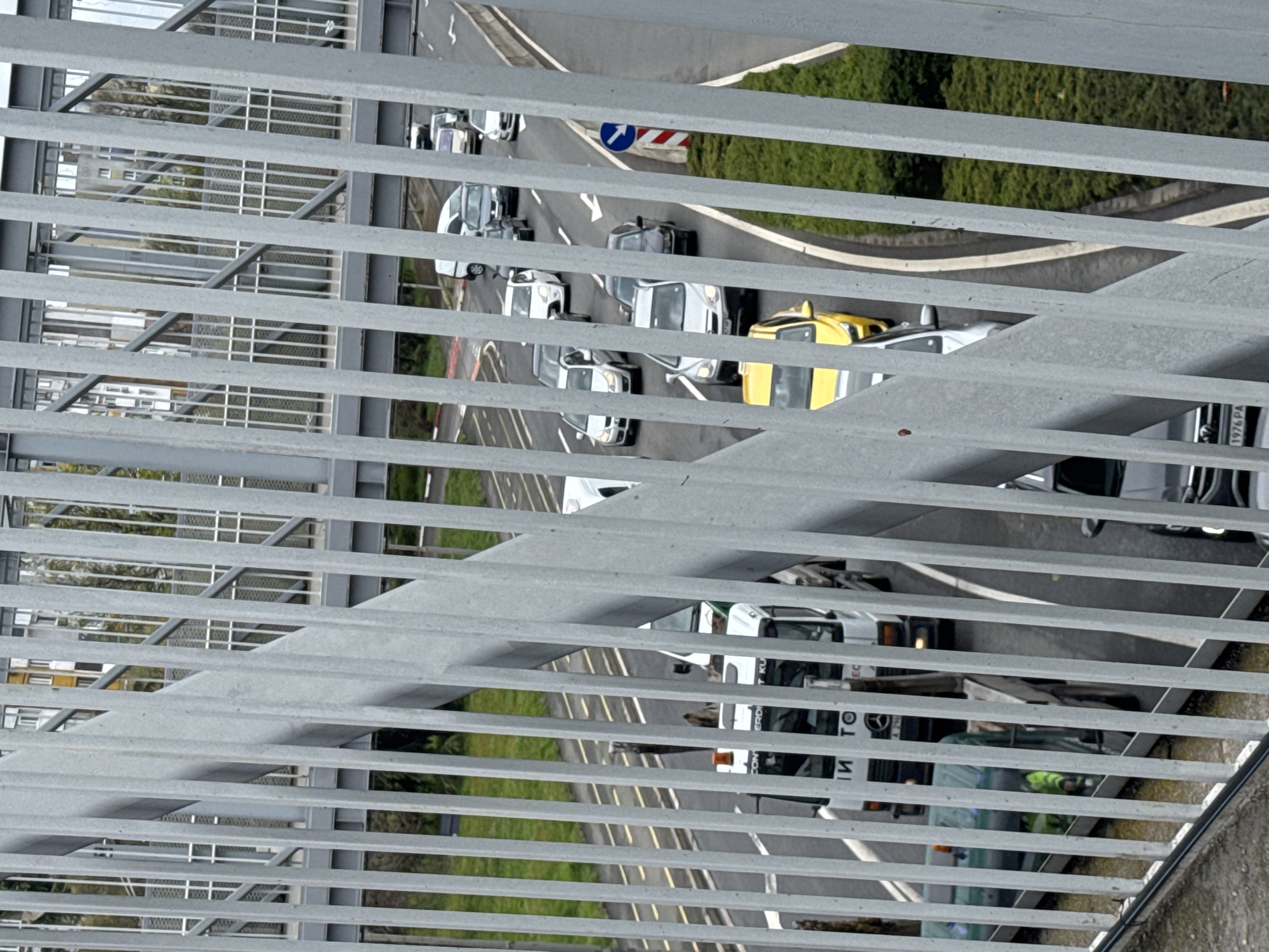 An elevated, angled view through a metal railing shows a multilane road with various vehicles. The vehicles include cars, vans, and trucks, with some people visible near a white truck. The environment includes green vegetation flanking the road. A sign with a blue arrow is partially visible. The general location is suggested as Burgas, Bulgaria.