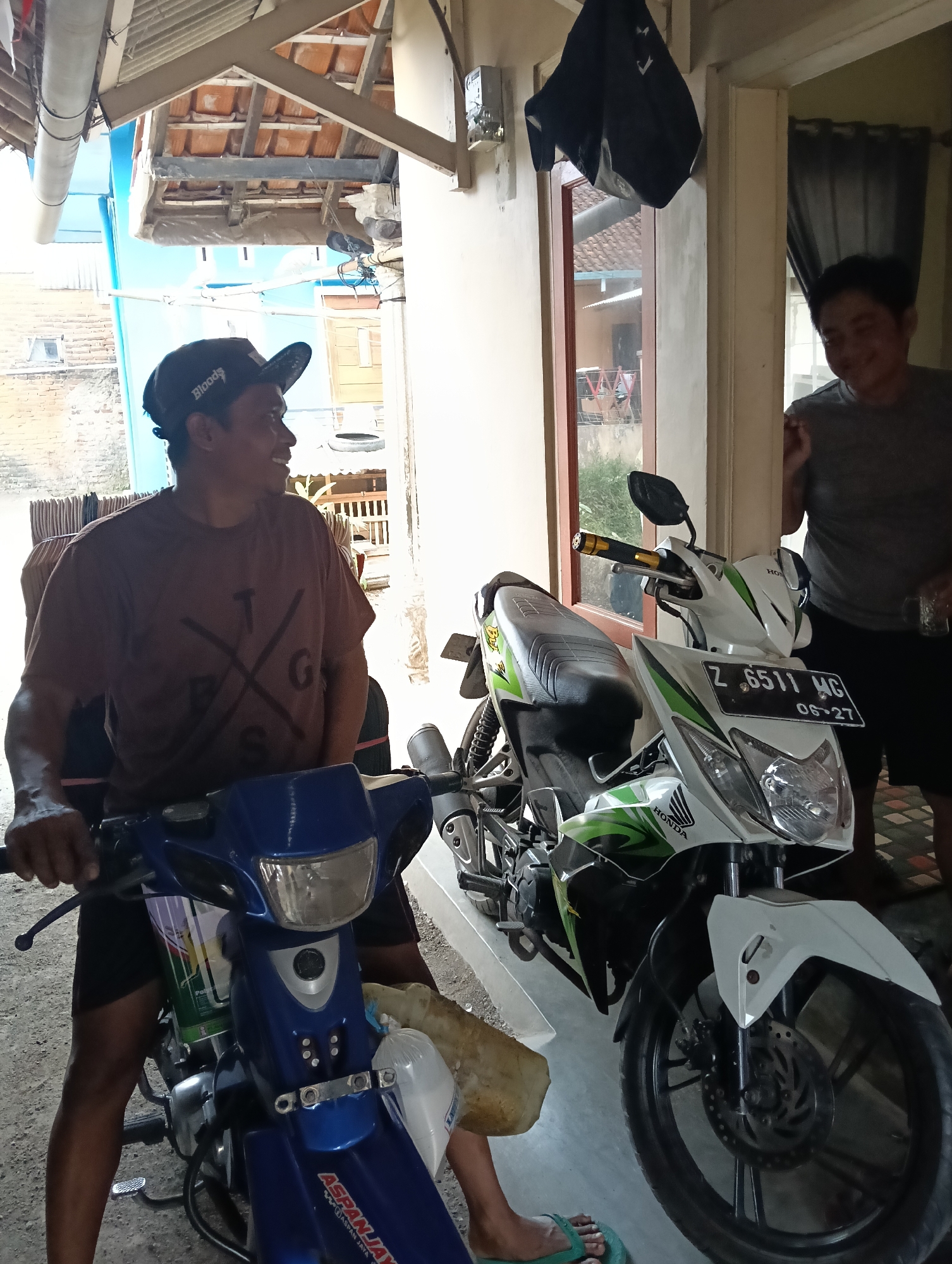 Two men are standing next to two motorcycles in what appears to be a residential area in Tasikmalaya, Indonesia. The man on the left is wearing a brown t-shirt and a black baseball cap and is seated on a blue motorcycle. He is looking to his right and smiling. The man on the right is wearing a gray t-shirt and black shorts and is standing behind a white and green motorcycle. He is looking at the man on the left and appears to be speaking. The motorcycles are parked in an outdoor area with a covered roof overhead. The environment is somewhat cluttered with various items, including what look like bundles of goods stacked behind the man on the left. The overall scene suggests a casual interaction or a moment of respite.