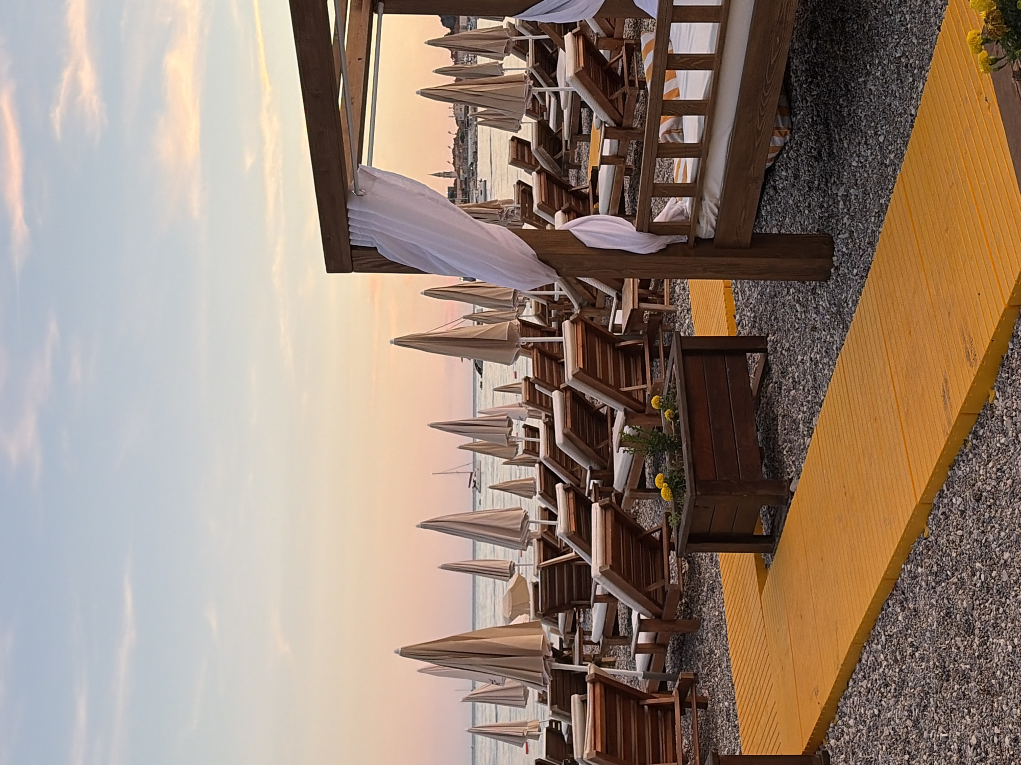This image captures a tranquil beach scene in Budva, Montenegro, during twilight. Rows of empty wooden lounge chairs and closed, beige umbrellas are arranged on a pebble-strewn beach, facing the calm sea. In the foreground, a bright yellow wooden walkway extends across the beach. To the right, a wooden cabana with white curtains and a daybed offers a shaded resting spot. In the distance, a sailboat glides across the water, and the silhouette of a coastal town with a prominent church spire is visible against the pastel-colored sky. The overall atmosphere is serene and inviting, hinting at a peaceful end to the day.