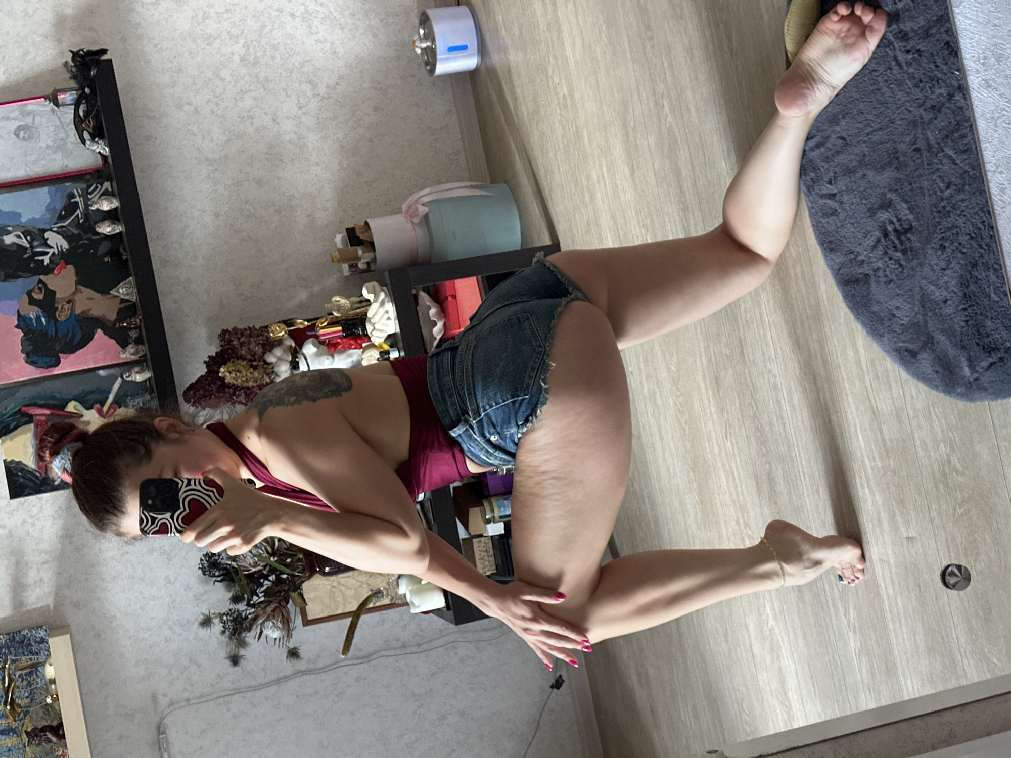 A woman is kneeling on a light-colored wooden floor, with her right leg extended and foot resting on a gray rug. She is wearing a red top and denim shorts. She is holding a smartphone in her left hand, presumably taking a photo of herself. The setting appears to be indoors, possibly a home. The background includes a shelf with various decorative items and artworks on the wall. There is no indication of the city of Vladimir, Russia.
