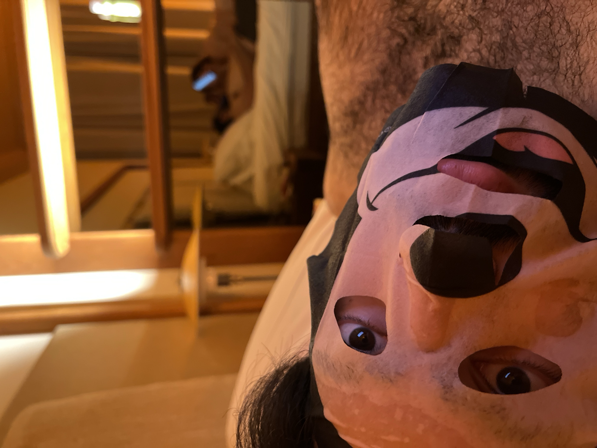 A person wearing a paper face mask is visible in close-up. Only the eyes, part of the nose, and the mask's design are visible. The mask has a black and pale peach color scheme. The background shows a room with wooden furniture and a person reflected in a mirror. A hairy chest is visible in the background, closer to the camera. The room has warm lighting. The location is unknown and could not be in Tel-Aviv, Israel.