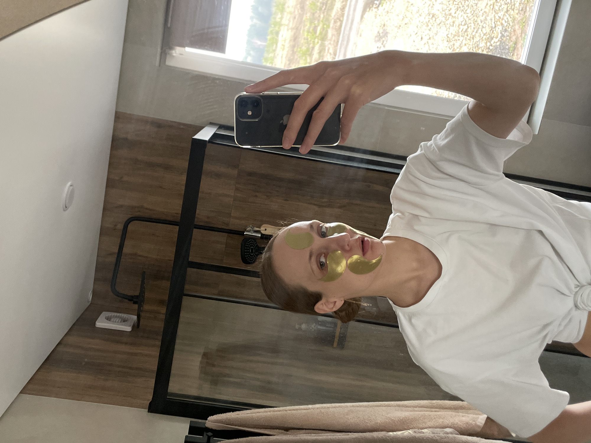 A woman is taking a selfie in a bathroom. She is wearing a white t-shirt and has gold eye masks on her face. She is holding a black smartphone in her left hand. The bathroom has wood-paneled walls and a window that looks out onto a field. The location is Podshivalovo, Russia. There is a shower with a black frame and a towel rack with towels on it.