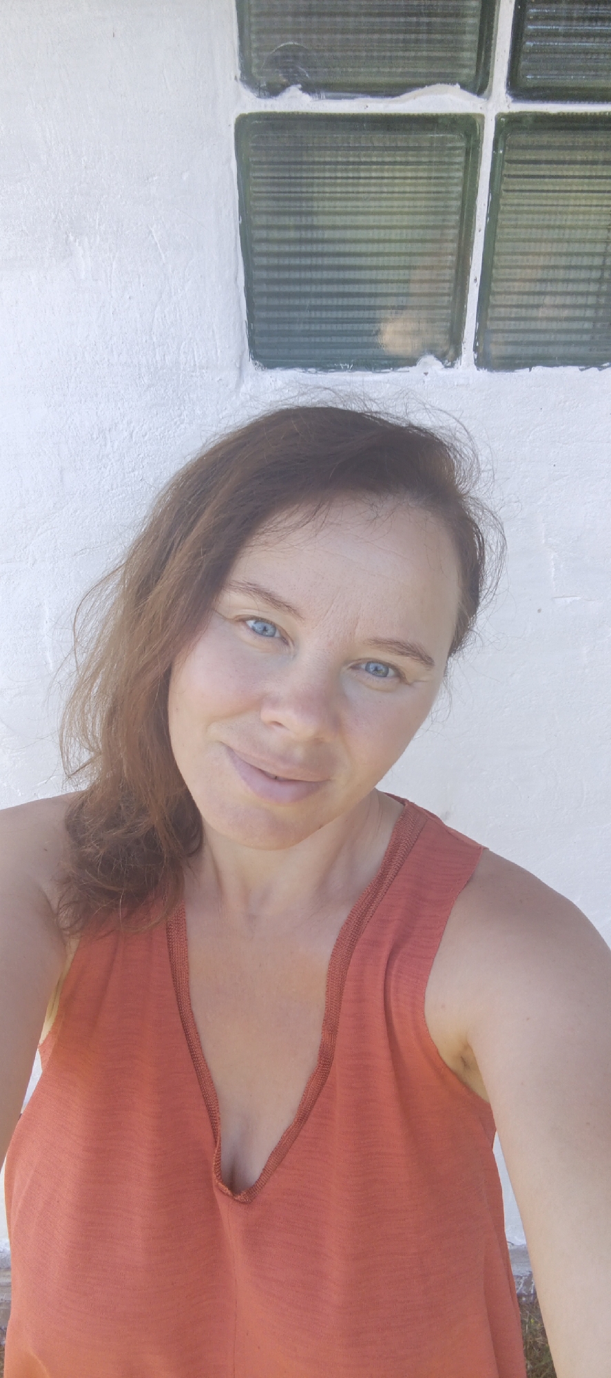 A woman with brown hair and blue eyes is taking a selfie. She is wearing an orange tank top. The background is a white wall with glass blocks in the upper right corner. The image appears to be taken outdoors on a sunny day. There is no identifiable location in the city of Chaikovsky, Russia.