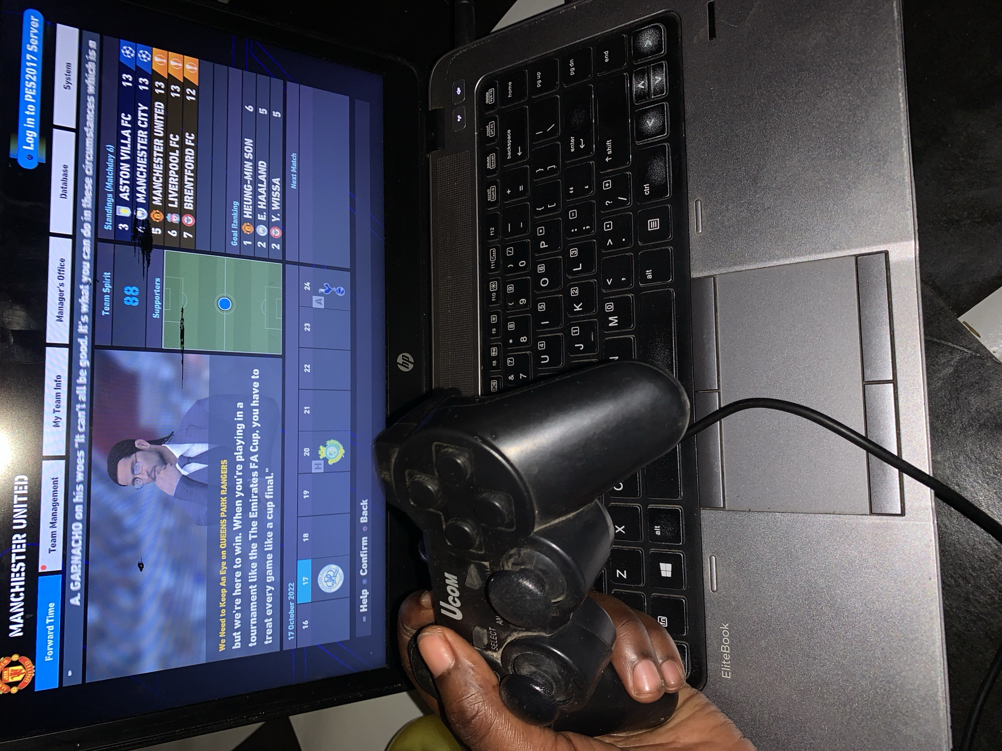 A person is holding a Ucom gamepad over a black and gray HP EliteBook laptop. The laptop screen displays a soccer game menu showing "Manchester United" on the left, a manager image, and standings. Aston Villa FC is at position 3, Manchester United at position 5 in the standings. Goal rankings on the right show Heung-Min Son at the top. The gamepad's cord rests near the laptop's trackpad. The environment is dimly lit.