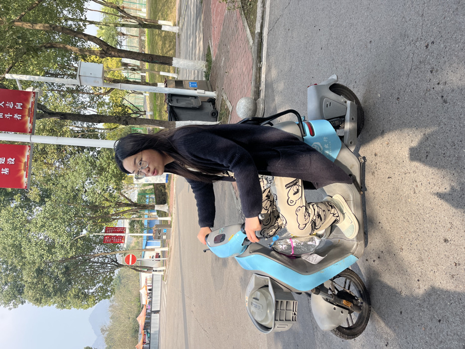 A young woman wearing glasses and a dark cardigan sits on a light blue electric scooter on a paved road. She is facing left and looking toward the camera. She is wearing pants with a cartoon bear pattern. The scooter is on a road with trees and buildings in the background. A red sign with Chinese characters is visible. The road is bordered by grass and trees on one side. The sky is overcast.