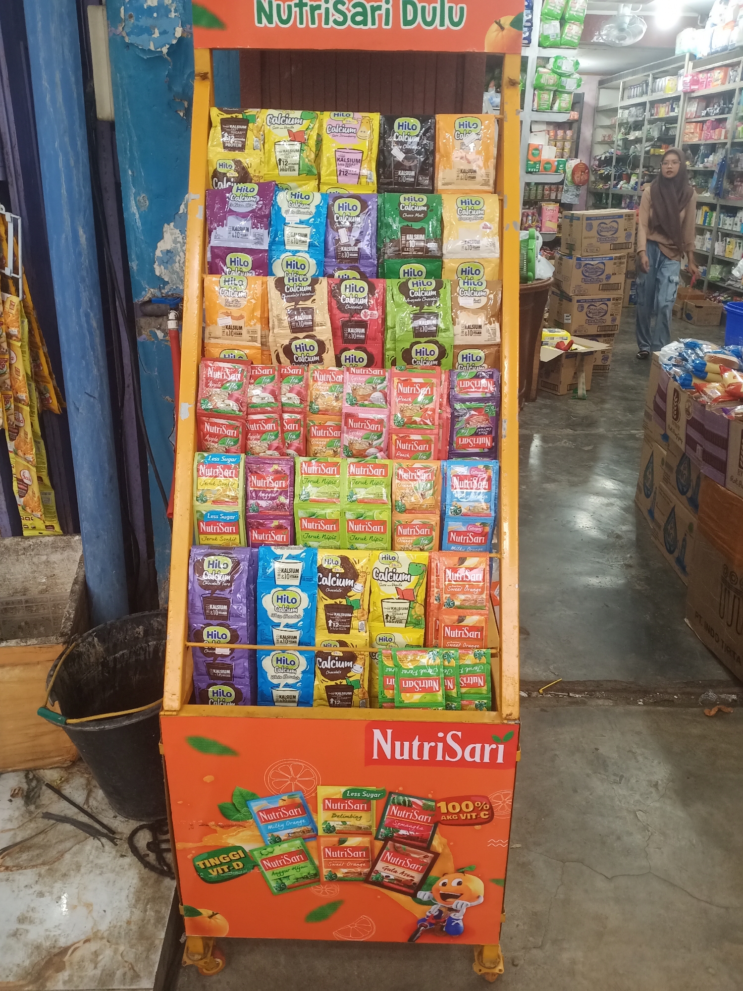 A NutriSari product display stand is positioned in front of a shop in Lhokseumawe, Indonesia. The stand is filled with various flavors of NutriSari and Hilo Calcium drink mixes, neatly organized in rows. The display features an orange-colored base with graphics of fruits and the NutriSari logo. To the right of the display, a woman wearing a hijab and a light brown top, along with light blue pants, walks past a stack of cardboard boxes. The background shows shelves stocked with other products, typical of a local convenience store or minimarket. The flooring appears to be concrete.