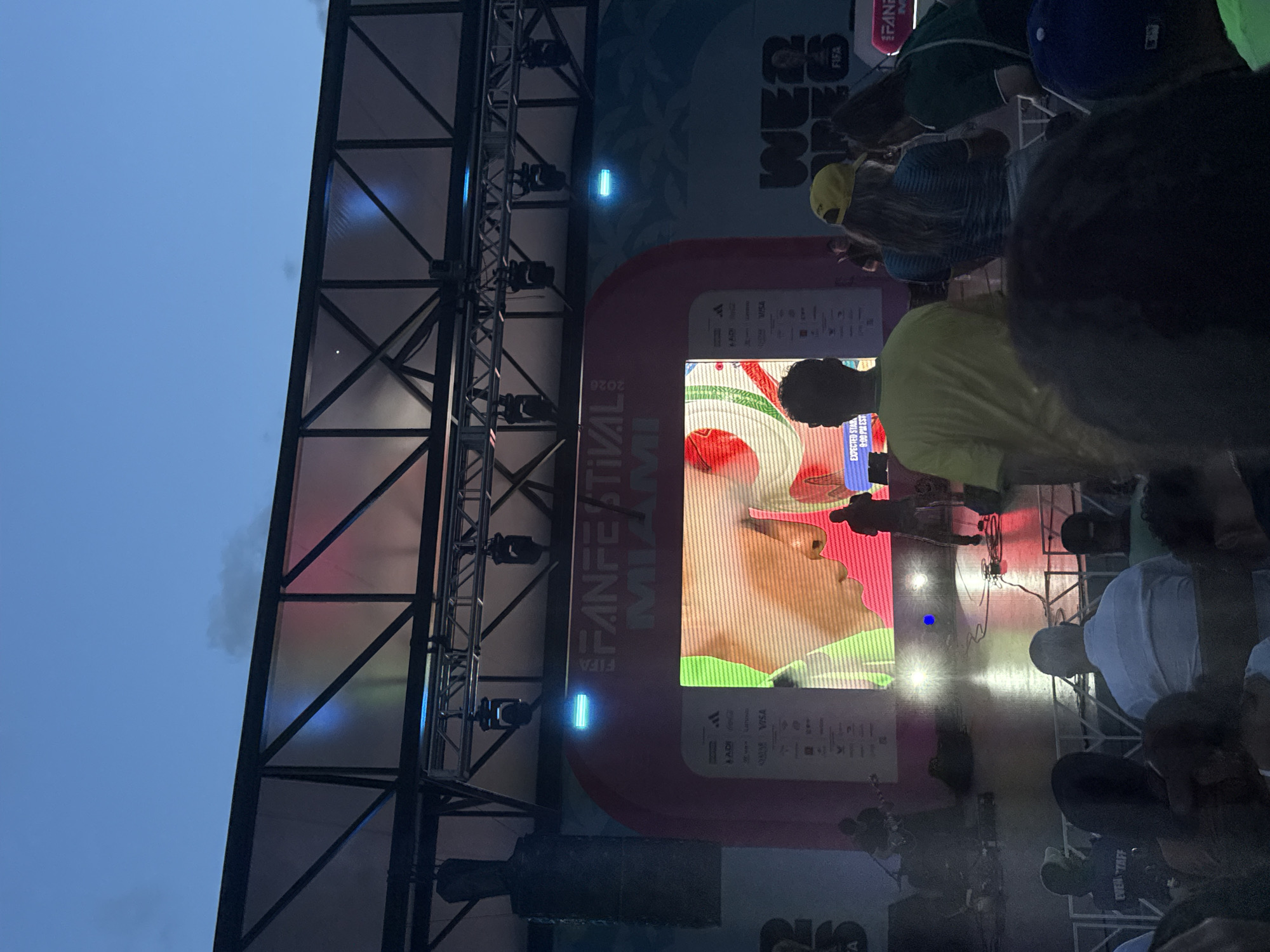 The image shows a crowd gathered at the FIFA Fan Festival in Miami, Florida. A large screen displays a close-up of a person's face, possibly a performer or athlete. The stage is lit with bright lights, and there is a band performing on the left side of the stage. The crowd is visible in the foreground, with some people standing and others sitting. The event appears to be taking place outdoors, with a clear sky visible above the stage. The overall atmosphere seems to be festive and energetic, with people enjoying the event.