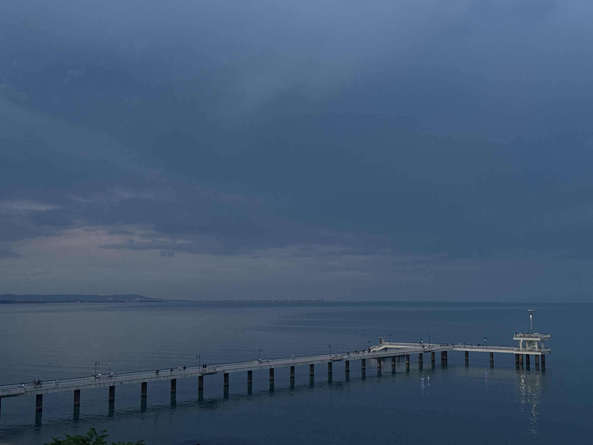 The image shows the Burgas Bridge in Burgas, Bulgaria, during twilight. The long, pier-like structure extends into the sea and culminates in a platform with a light fixture. People are sparsely distributed along the bridge. The sky is overcast, and the sea is calm. In the distance, a coastline with buildings is visible. The overall tone is subdued due to the time of day.