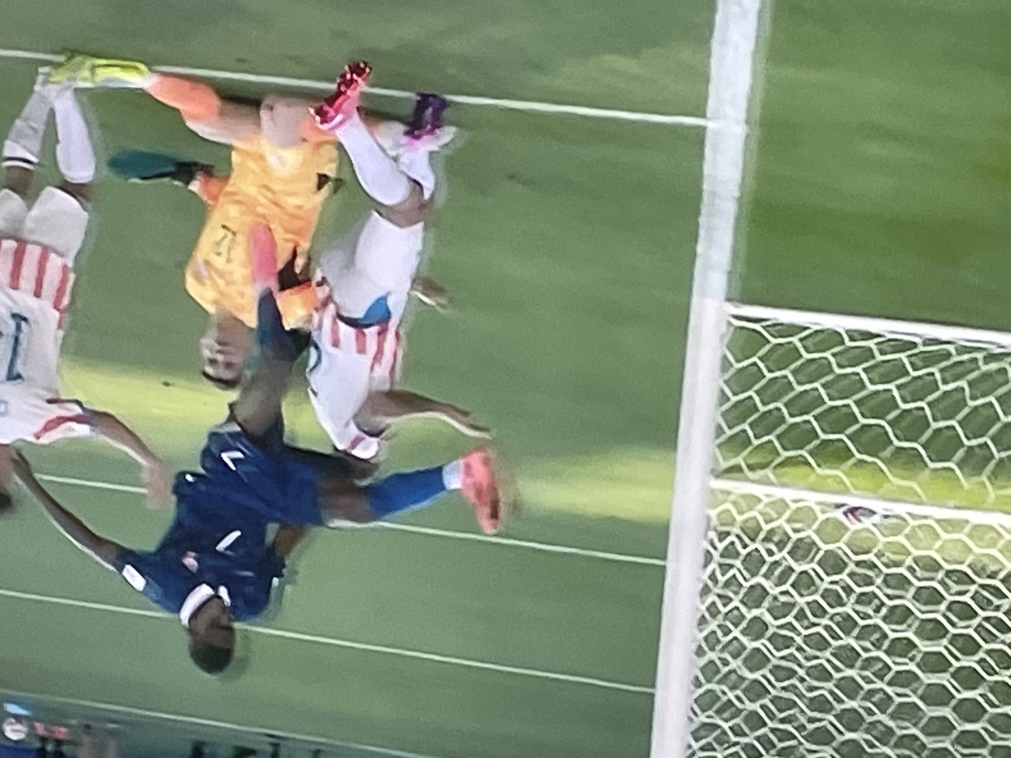 In Zhukovo, Russia, a soccer game is underway. In the foreground, a player in a blue uniform with the number 7 on his jersey is in mid-air, kicking a soccer ball. He is wearing bright orange cleats. Below him, a player in a white jersey with red stripes and orange shorts is on the ground, also in action. The goalkeeper, wearing an orange jersey with the number 12 and black gloves, is on the ground, reaching for the ball. Another player in a white jersey with red stripes and white shorts stands to the right. A soccer net is visible in the background. The game is being played on a green grass field with white markings.