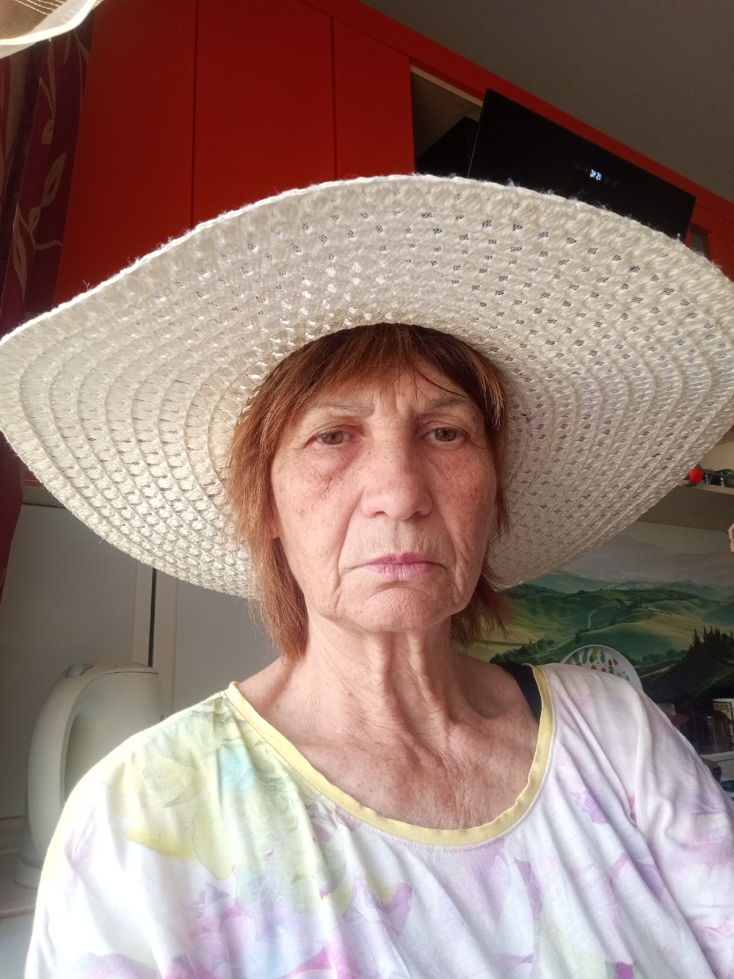 A medium shot shows an older woman facing forward, wearing a wide-brimmed white woven hat. She has reddish-brown hair and is wearing a light-colored, loosely fitting top with a floral pattern in shades of pink, yellow, and green. The background appears to be indoors, with a bright orange surface on the left and a dark object, possibly a kitchen appliance or electronic device, on the upper right. A painting of a green, hilly landscape is visible behind the woman. The lighting suggests it is daytime. The woman's expression is neutral. The image does not contain any information that would suggest a specific location within the city of Mykolaiv, Ukraine.