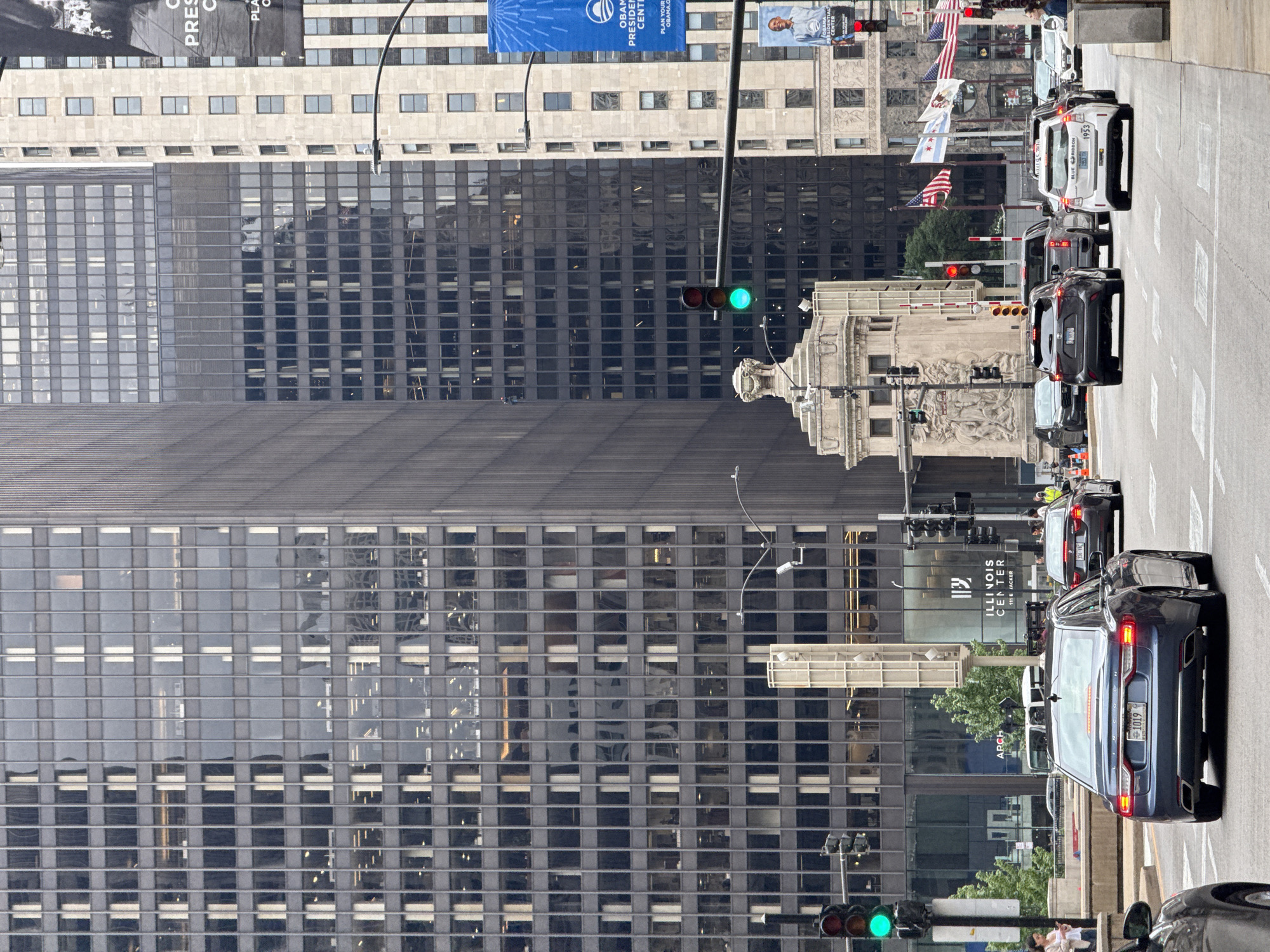 This media file shows a street scene in Chicago, USA, with tall, modern buildings. The buildings feature dark glass facades with grid-like window patterns, reflecting the sky and surrounding structures. In the foreground, a dark blue Lincoln sedan is parked on the right side of the road, with its license plate displaying "1019". Other vehicles, including a white van with "BLUE RIBBON 1953" visible on its side, are also present in traffic.
American flags and an Illinois state flag are seen flying on poles attached to the buildings. Banners with text like "OBAMA PRESIDENTIAL CENTER" and "PLAN YOUR OBAMA.C" are also displayed. The scene appears to be during the daytime, under overcast or slightly cloudy skies, as indicated by the muted lighting and reflections. Pedestrians are visible on the sidewalk near the bottom right of the image. Traffic lights are also present, with one showing a green signal. The overall impression is a bustling urban environment with a mix of architectural styles and civic symbols.