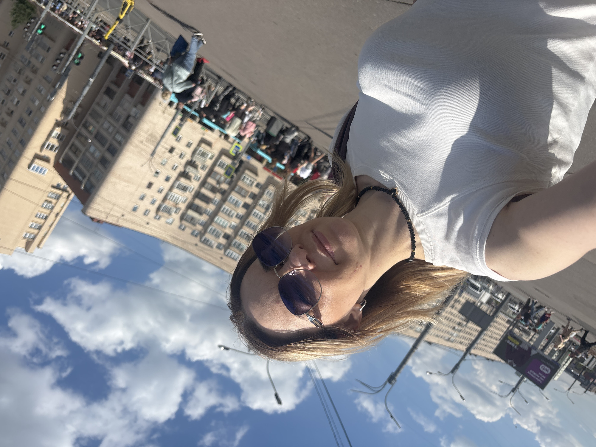 A low-angle, rotated selfie captures a person in the foreground against a vibrant city backdrop in Saint Petersburg, Russia. The main subject, likely female, has blonde hair and is wearing round sunglasses and a white tank top. She sports a dark, beaded necklace and displays a relaxed expression with a subtle smile directed towards the camera.
The background reveals a bustling urban street scene under a clear blue sky dotted with fluffy white clouds. Tall, multi-story buildings, characteristic of city architecture, line the street. Several streetlights and overhead wires traverse the scene, with a traffic light showing a green signal visible on the far left. Numerous blurred figures populate the street and sidewalks, suggesting pedestrian and vehicle activity. One person in the mid-ground appears to be on a scooter or similar device. An advertising board is visible in the middle-right, showing partially obscured text that includes "Q&M" and some blurry Cyrillic characters.
The bright sunlight indicates a daytime setting with pleasant, clear weather. The overall atmosphere is casual and lively, typical of a city on a sunny day.