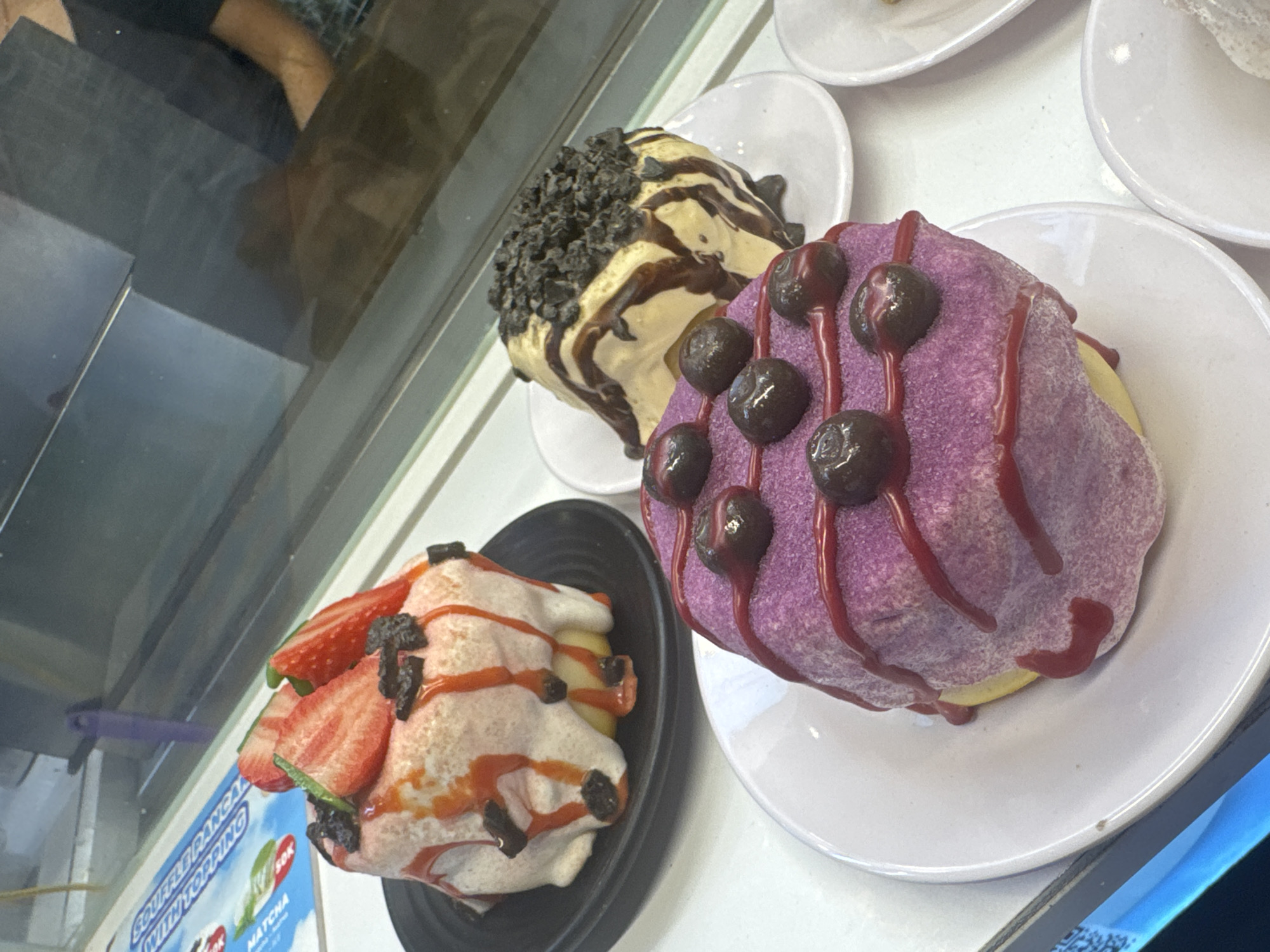 The image captures a display case featuring three plates of decorative food items, likely desserts. The bottom plate holds a pastry topped with white cream, sliced strawberries, and dark drizzle or pieces. Next to it, on a higher level, is another plate with a beige pastry decorated with chocolate chips and a dark-colored drizzle. The top plate contains a purple pastry with dark round toppings and a red drizzle. The items are positioned to be viewed from the front, indicating a customer-facing display, potentially in a bakery or dessert shop.