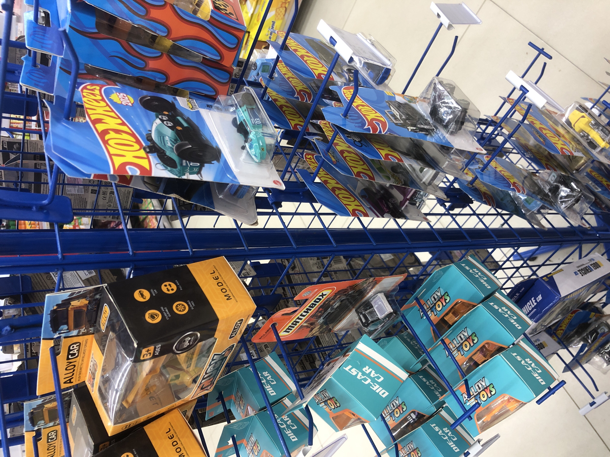 The image shows a metal blue rack displaying various toy cars for sale. The rack is filled with blister packs and boxed toy cars. Brands visible include Hot Wheels, Matchbox, and Alloy Cars. The toy cars are packaged in colorful cardboard and plastic. The background is blurred but appears to be an indoor retail environment with shelves stocked with other products. Based on the context provided by the prompt, this scene is located in Langsa, Indonesia.