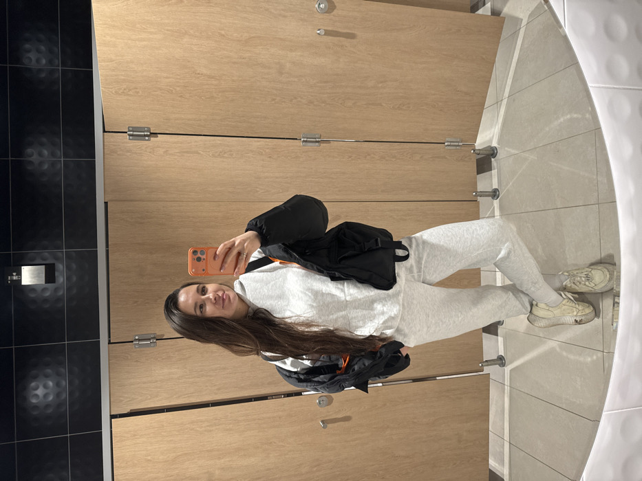 A woman takes a selfie in a bathroom with light brown wooden stalls and tiled walls. She wears a light grey sweat suit, white sneakers, and a black jacket. She holds an orange phone in her right hand, angled towards the mirror. Her long brown hair cascades over her shoulder. The bathroom features a combination of black and light grey tiles on the walls and floor.