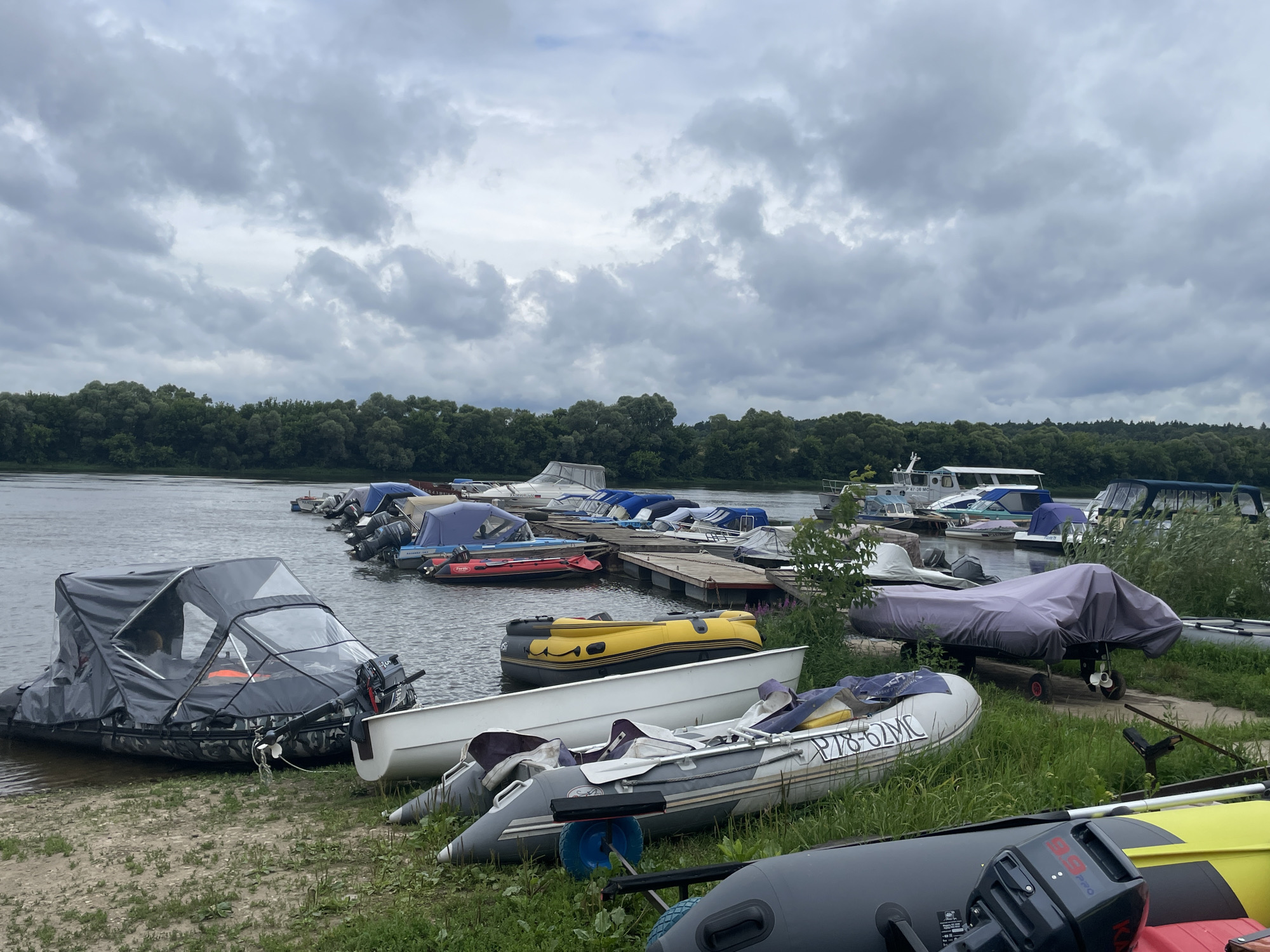 An outdoor, ground-level shot captures a scene by a river, likely in Pushchino, Russia, under an overcast sky. The foreground is occupied by several inflatable boats of various sizes and colors, some resting on the grassy bank and others partially in the water. One prominent inflatable boat in the foreground is covered with a grey canopy and has a dark camouflage pattern on its sides. Next to it, a white boat lies on the shore, followed by a grey inflatable boat with white accents, and further back, a yellow inflatable boat. In the middle ground, a wooden dock extends into the river, lined with numerous other boats of different types, many of which are covered. The river stretches into the background, with a dense line of green trees bordering the opposite bank. The sky is filled with heavy, grey clouds, suggesting impending rain or a generally gloomy day. No people are actively engaged in any activity, though the presence of the boats implies a recreational or logistical purpose related to the river.