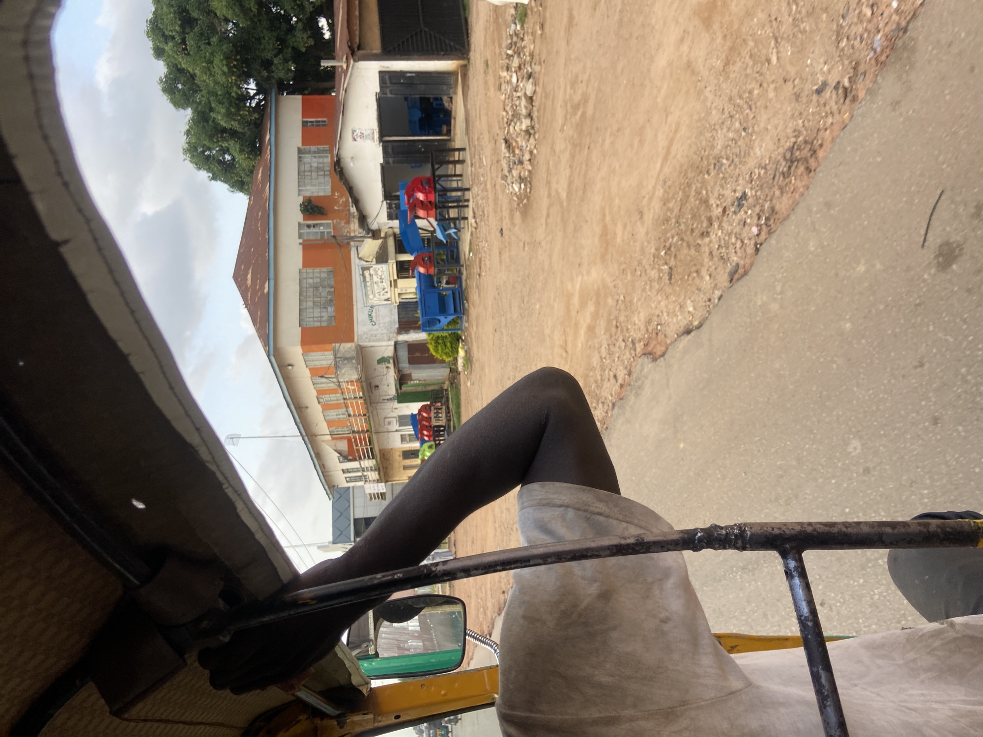 The image depicts a scene from the perspective of someone riding in a three-wheeled vehicle, likely a tuk-tuk, in Jos, Nigeria. The vehicle's interior, including the roof lining and part of a metal bar, is visible in the foreground. A person's leg, clad in dark leggings, and torso, wearing a light-colored garment, are also prominent. The scene outside shows a dusty, unpaved road next to a paved surface.
Buildings with orange and white facades line the road, some appearing to be shops or homes. Several blue and red pieces of equipment, possibly industrial machinery or seating, are displayed outside one of the buildings. A large tree with green foliage is situated to the left, suggesting a somewhat rural or semi-urban environment. The sky is overcast with clouds. The overall impression is a snapshot of daily life in a Nigerian town, capturing the movement and streetscape. The time of day appears to be daytime, with the overcast sky suggesting potentially cloudy weather. There is no discernible text visible in the image.