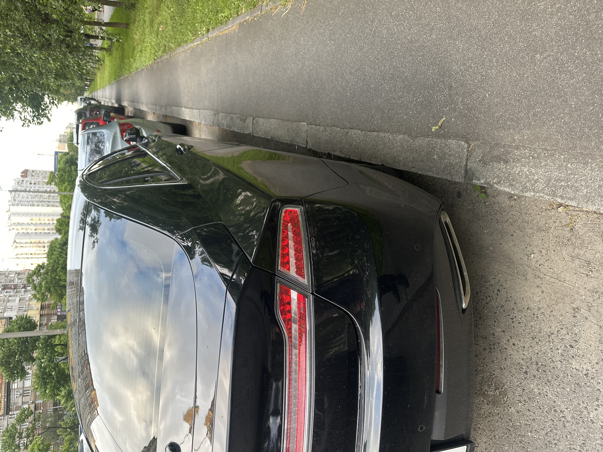 In Kyiv, Ukraine, a black sedan is parked on the side of a street adjacent to a sidewalk lined with trees. The car is viewed from the rear three-quarter angle, showing its taillight and exhaust pipe. In the background, apartment buildings and more trees are visible, suggesting a residential or mixed-use urban area. The sidewalk appears to be made of asphalt with a concrete curb. Other cars are parked in a line behind the black sedan, and a person is walking on the far side of the sidewalk.