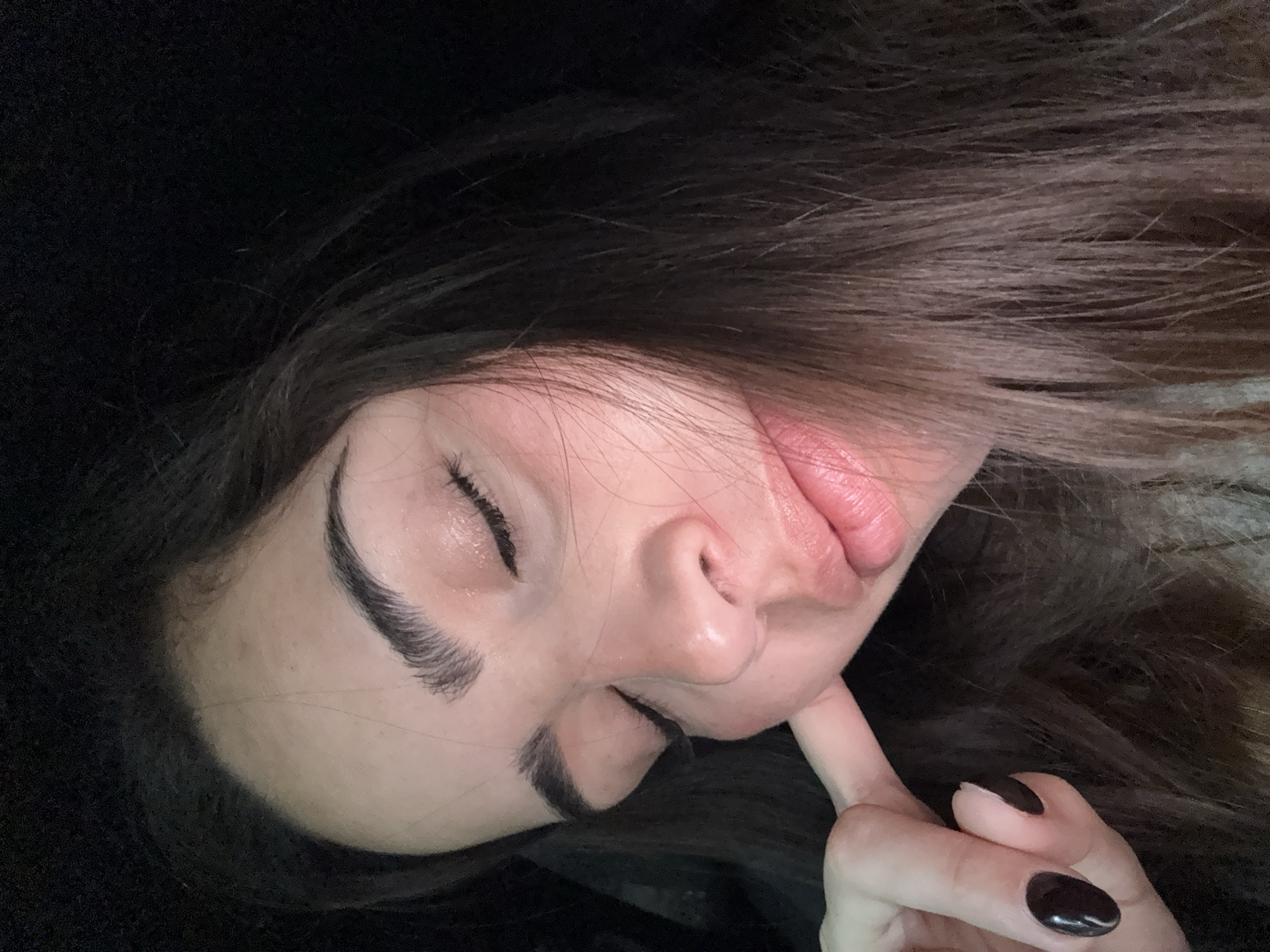 A woman is lying down with her eyes closed and her index finger touching her chin. She has dark hair and dark eyebrows. Her nails are painted dark. The image is taken at close range and is rotated counter-clockwise. The location in San Bernardino Tlaxcalancingo, Mexico, is not determinable from this image.