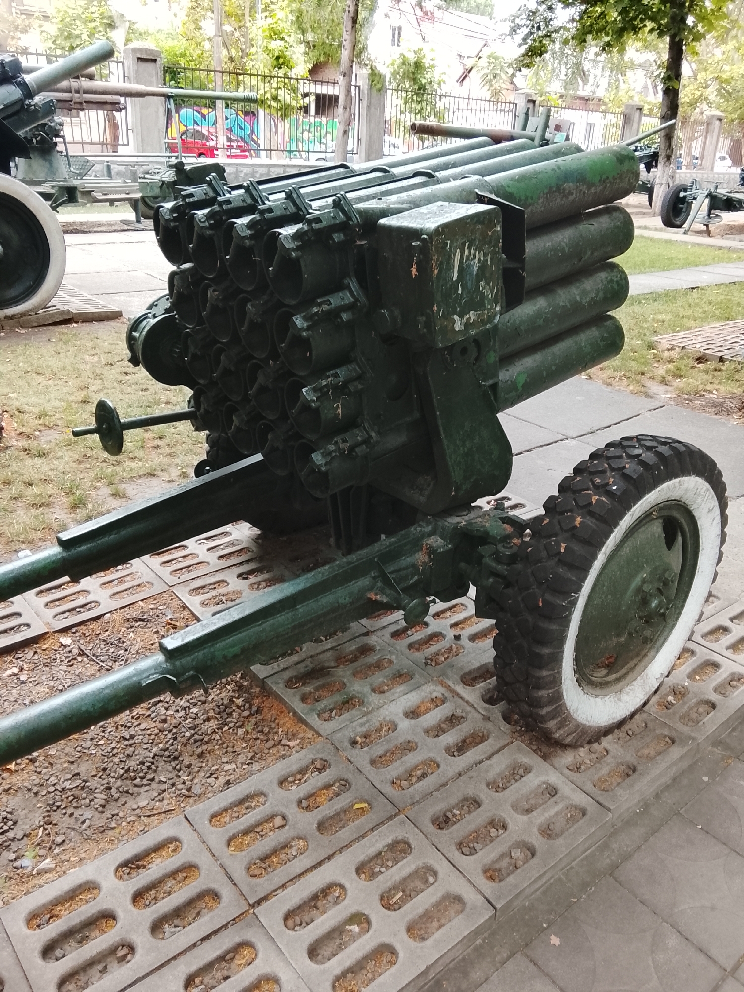 The image shows a military exhibit outdoors, likely in Chișinău, Moldova, given the style of the fence and the building in the background. In the foreground, a dark green, multi-barreled rocket launcher is positioned on a paved area constructed with large, rectangular concrete blocks with oval cutouts, interspersed with gravel. The launcher has a large, robust frame with two main support arms extending forward, one of which is attached to a spoked wheel with a white-rimmed, knobby tire. The rocket tubes are arranged in multiple rows and columns, appearing to be a significant part of the weapon. The metal surfaces of the launcher show signs of wear and some greenish-gray paint. Behind the rocket launcher, a different artillery piece with a long barrel and a wheel is partially visible. Further in the background, a black metal fence with decorative finials encloses an area with trees and some buildings. Graffiti with bright colors is visible on a wall behind the fence. The lighting suggests it is daytime, with overcast conditions. There are no people or animals present in the image.