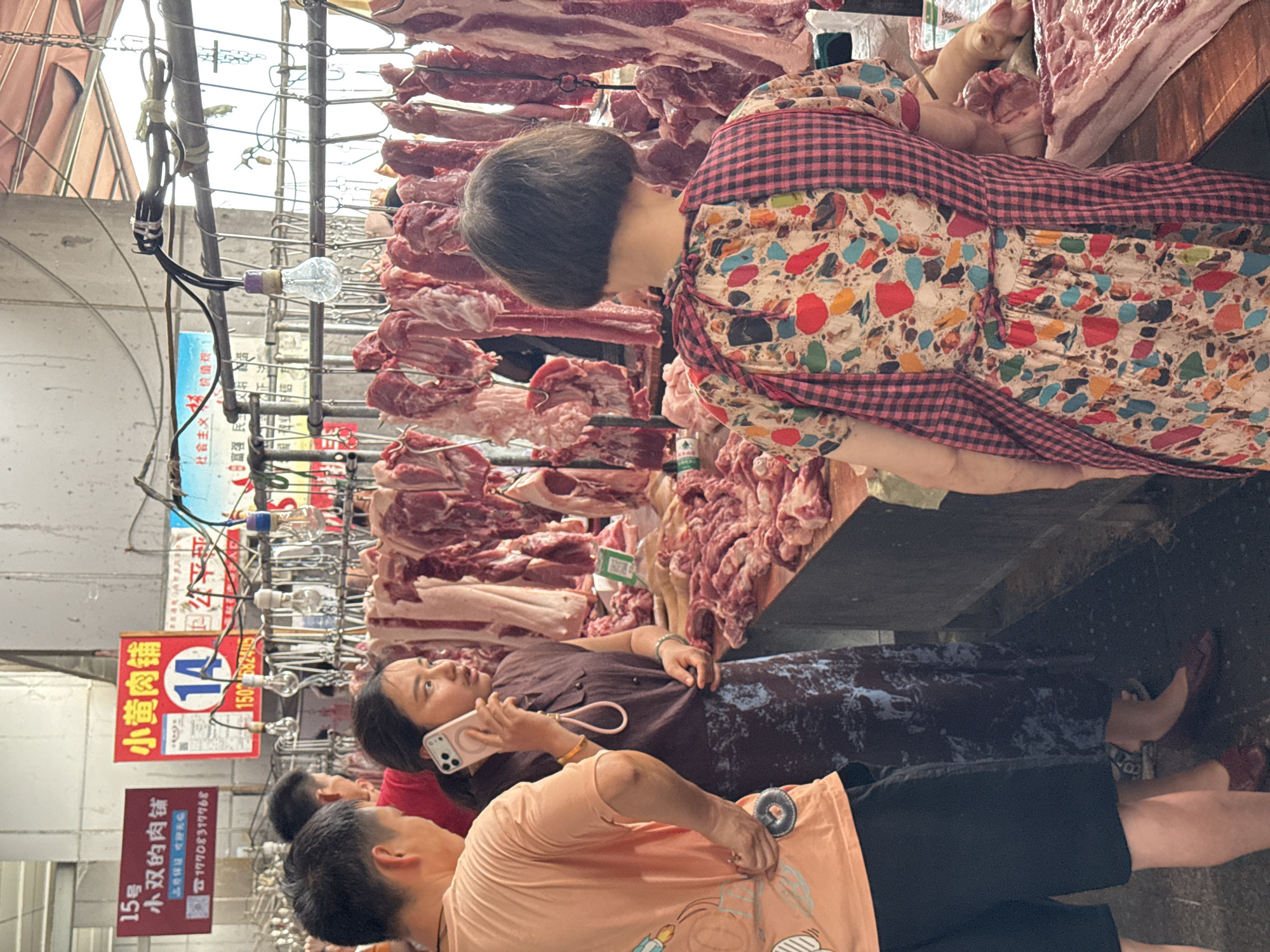 This image depicts a bustling indoor meat market in Jiangjin, China. The scene focuses on a meat stall featuring numerous cuts of raw pork, displayed hanging from metal racks and piled on a wooden counter in the foreground. Three individuals are prominently featured. On the left, a man wearing an orange t-shirt with a graphic design and dark shorts stands with his back partially to the viewer, observing the stall. In the center, a woman with dark hair tied back wears a dark purple dress and holds a light-colored smartphone to her right ear, appearing to be on a call while looking towards the right side of the frame. Her left hand is near her waist. On the right, a woman with a short, dark bob haircut is seen from behind, wearing a colorful, abstract-patterned dress overlaid with a pink and white checkered apron tied at the back. She is positioned directly in front of the meat counter, facing the displayed products.
In the background, behind the people, multiple large cuts of meat hang from hooks on a multi-tiered metal rack structure. Above the stall, various signs are visible. A red rectangular sign on the left reads "15号 小双的肉铺" (No. 15, Xiao Shuang's Meat Shop) with a phone number "17708317768." Adjacent to it, a larger red sign reads "小黄肉铺" (Little Huang's Meat Shop), featuring the number "14" in a blue circle and a phone number "1502562402." Exposed wiring and bare light bulbs hang from overhead structures, providing illumination. The overall environment appears functional and utilitarian, typical of a busy market setting.
