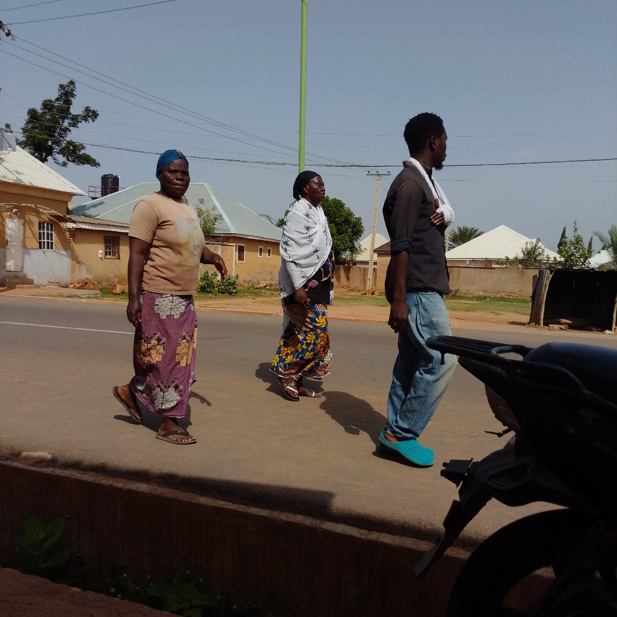 Three people are walking on a paved road in Gombe, Nigeria. The person on the left is a woman wearing a light brown t-shirt and a purple patterned skirt. She is wearing flip-flops and has a blue headscarf. The woman in the middle is wearing a white patterned scarf over a dark top and a colorful patterned skirt, also wearing flip-flops. The person on the right is a man wearing a dark shirt and blue jeans. He has a white bandage wrapped around his left arm and is wearing turquoise slip-on shoes. A portion of a black motorcycle is visible in the foreground on the right. The background shows residential buildings with light-colored roofs, trees, and utility poles with power lines. The sky is clear and blue. The scene appears to be a sunny day in an urban or semi-urban area.