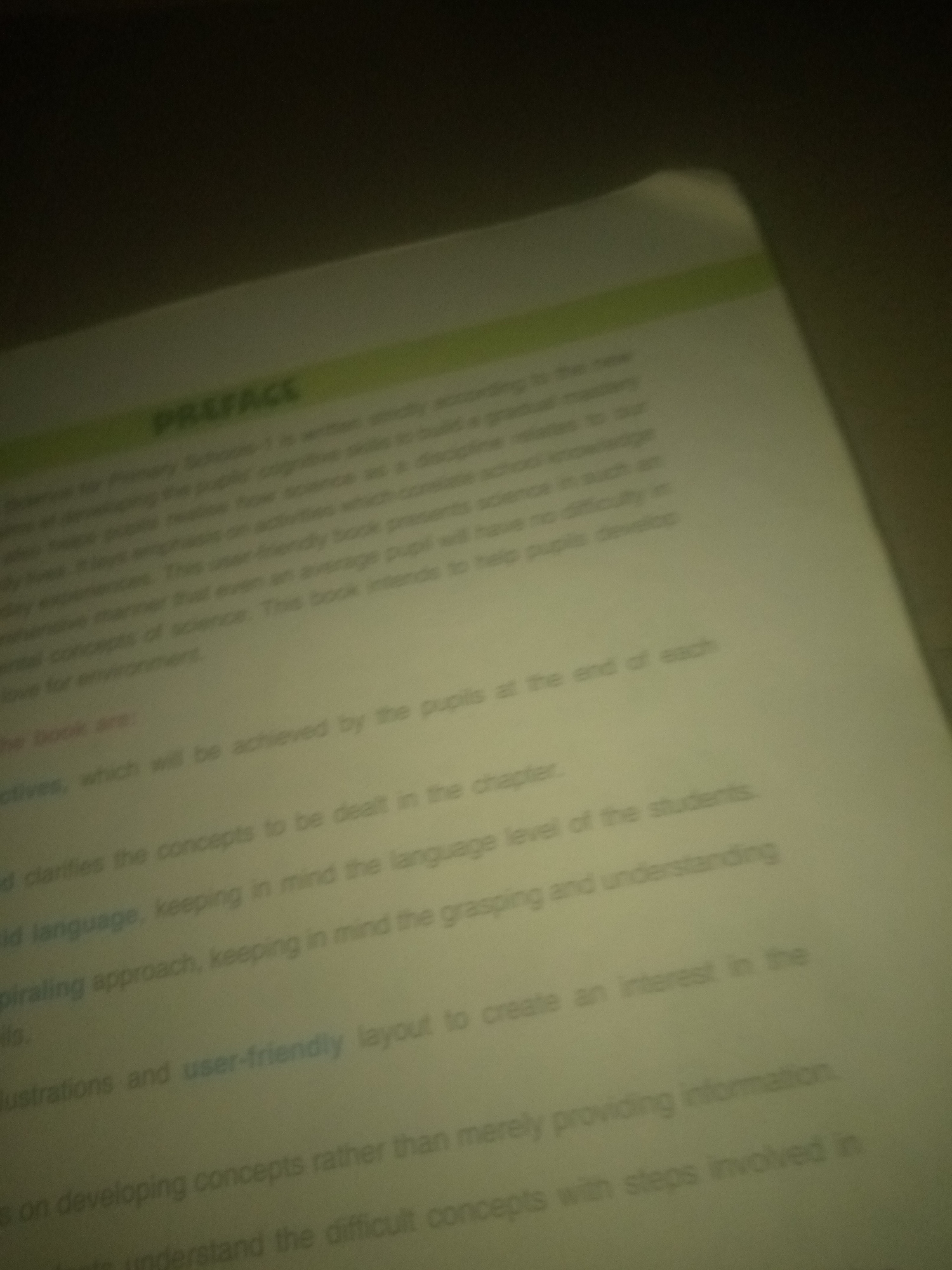 The image is a close-up, slightly blurry, overhead shot of an open book with a green strip running across the top of the page. The text is in English and appears to be a preface or introduction to a textbook. The main title "PREFACE" is visible in large, bold letters. Below the title, there are several paragraphs of text that discuss the book's approach to teaching science, emphasizing user-friendliness, age-appropriateness, and fostering interest in the subject. Specific phrases like "cognitive skills," "gradual mastery," "user-friendly book," "language level of the students," and "spiraling approach" are discernible. The background is dark and out of focus, suggesting the image was taken in low light conditions. There are no people, specific locations, or environmental details beyond what is visible on the printed page.