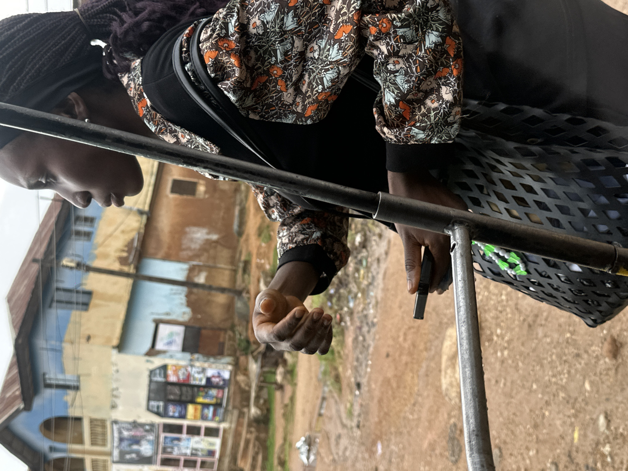 A medium shot captures a young Black woman in profile, facing right, holding a mobile phone in her left hand. She wears a black headscarf and a patterned blouse with black accents. Her dark braided hair cascades down her back. She is outdoors, with a blurred background of buildings and a dirt road. A metal railing runs horizontally across the foreground, and a black mesh bag hangs from her shoulder. The scene appears to be in Gwong, Nigeria.