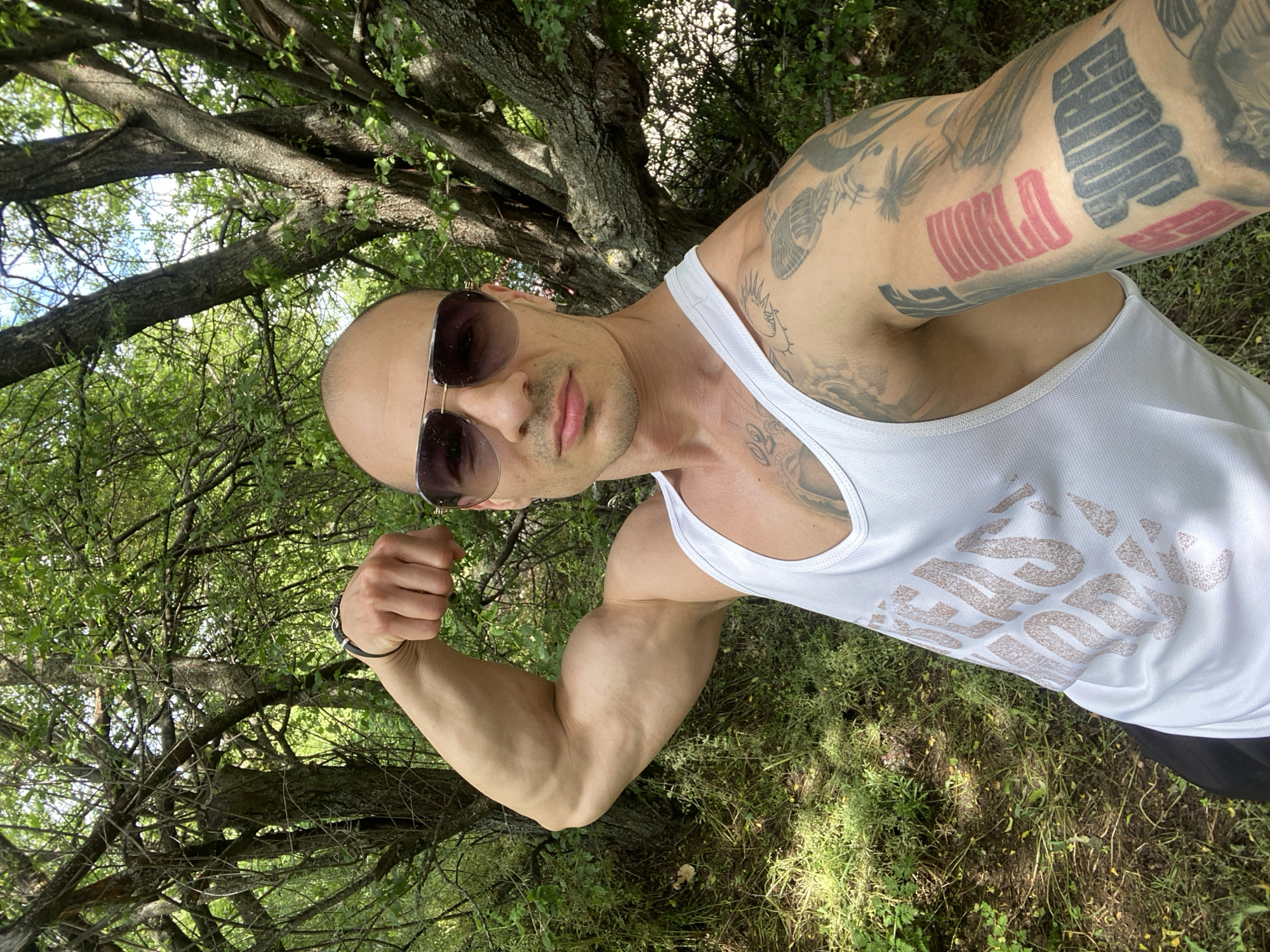 A bald man wearing sunglasses and a white tank top with a tan-colored graphic is flexing his right bicep. He has multiple tattoos on his arms and chest. The background consists of trees and green foliage. The location appears to be outdoors, possibly a park or wooded area in Pleven, Bulgaria.